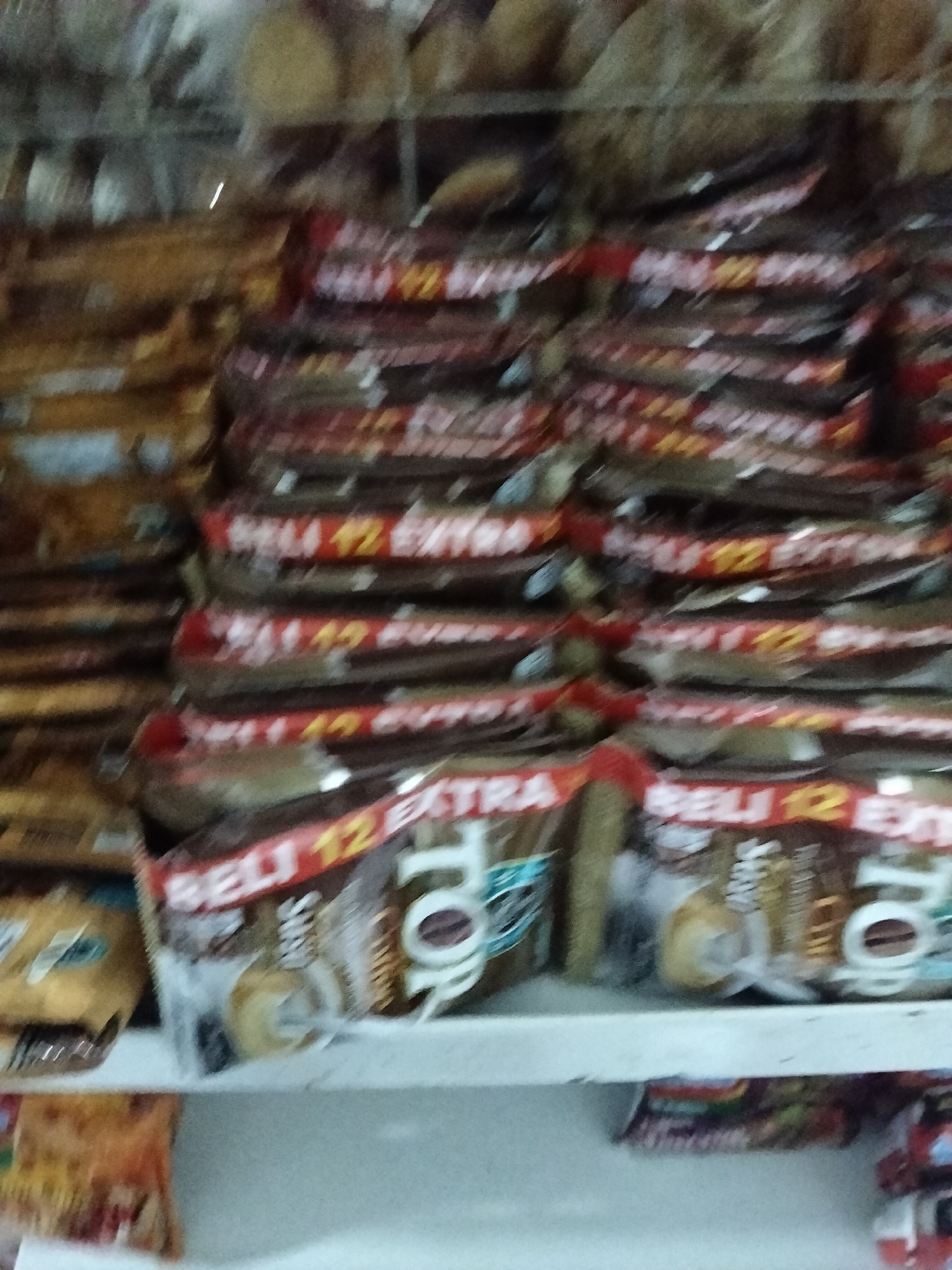 A shelf of packaged coffee products is displayed in a store in Gempolan, Indonesia. The products are stacked horizontally in rows, with the majority of them featuring red and white packaging. Some packages have text that reads "BELI 12 EXTRA" and "TOP COFFEE". The lighting in the store is dim, and the image is blurry, making it difficult to discern finer details. There are no people visible in the image. The shelf appears to be made of white material and is part of a larger retail display.