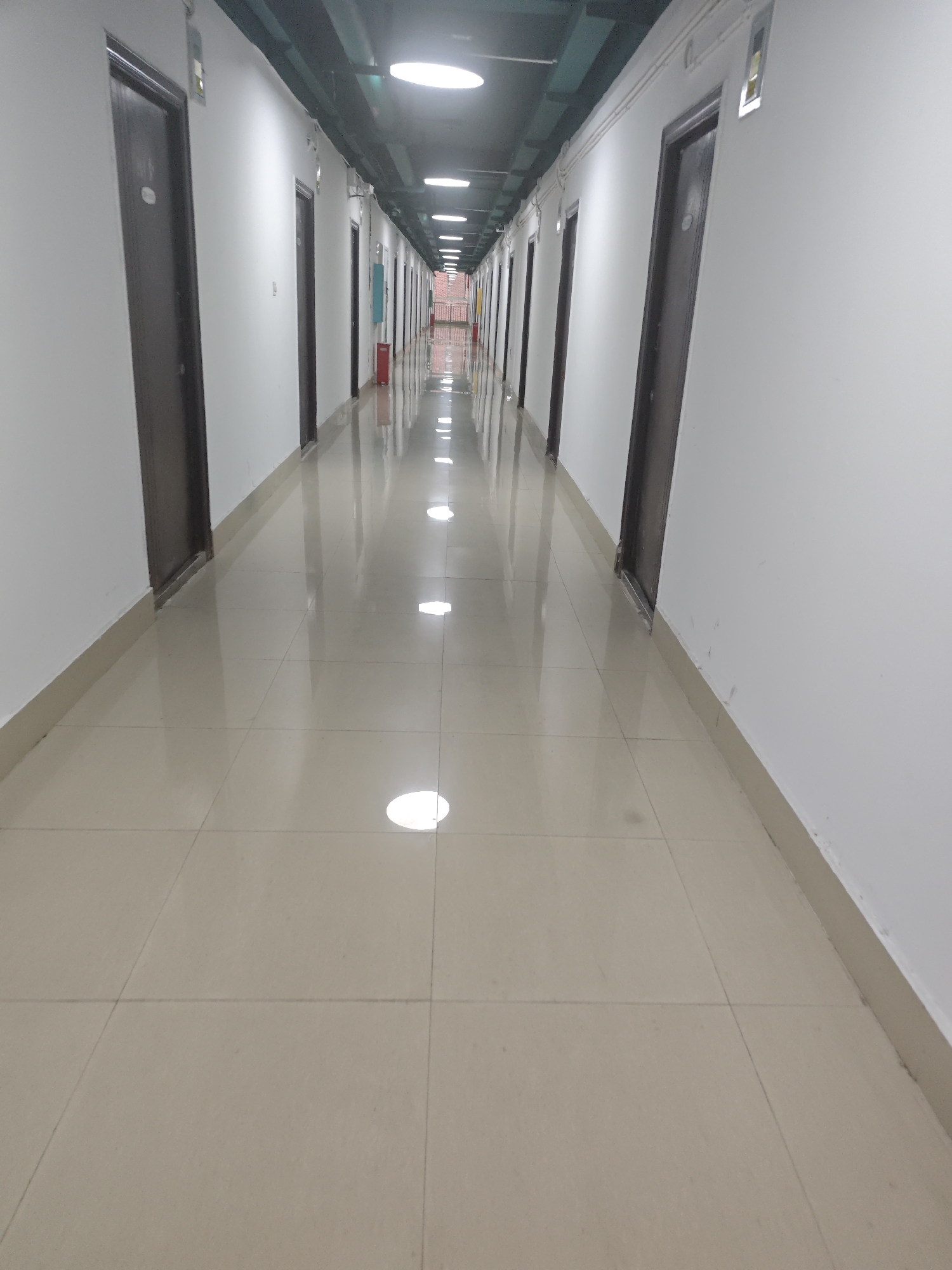 The image shows a long, well-lit corridor with doors lining one side. The corridor has a polished, reflective floor, suggesting it is clean and well-maintained. Fluorescent lights are visible in the ceiling, providing ample illumination. There are no people or any other living beings visible in the scene. The environment appears to be a modern building, possibly a hotel, dormitory, or office building, located in the Chongqing High-tech Zone, China. The doors appear to be uniform in design, each with a number plate or label. The walls are painted white, contributing to the bright and clean aesthetic of the space. The perspective of the image draws the viewer's eye down the length of the corridor, creating a sense of depth and order.