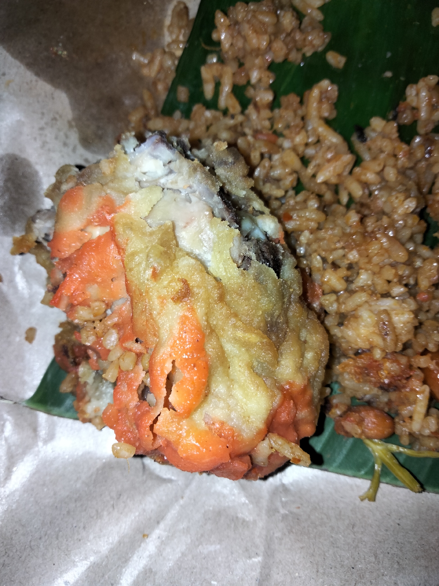 The image shows a close-up of a portion of food, likely served in a casual setting. The primary subject is a piece of battered and fried food, appearing to be fish or seafood, with a distinct orange hue from what could be a sauce or marinade, and a golden-brown batter. This battered item is partially resting on a bed of rice. The rice is cooked and appears to be fried, with a reddish-brown color, suggesting it's seasoned or mixed with ingredients. The food is presented on a large green leaf, which is a common practice for serving food in many parts of Indonesia, including Payagaboh. The leaf itself is placed on what looks like brown paper wrapping, typical of street food or takeaway packaging.
The lighting suggests it might be indoors or under artificial light, as there are no strong shadows that would indicate direct sunlight, nor is there any clear indication of the time of day. The background is blurred, focusing attention on the food. The texture of the battered item is rough and crispy from frying, while the rice grains are distinct and slightly glossy. A few wisps of green, possibly herbs or garnish, are visible near the edge of the food. The image composition is tight, emphasizing the details of the food itself. There is no visible text in the image. The overall impression is of a delicious and perhaps savory dish, prepared and presented in a traditional manner in Payagaboh, Indonesia.