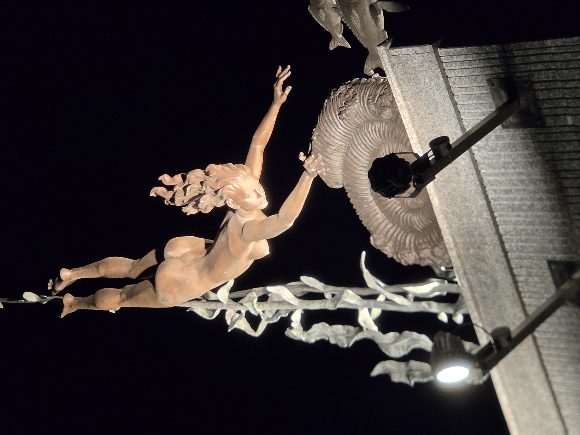 This is a night-time outdoor photograph of a bronze sculpture depicting a female figure in mid-air, suspended by an unseen element from above, with her body arched backward. The sculpture appears to be part of a larger installation that includes stylized representations of aquatic elements like seaweed and possibly fish, as well as a large, spiral-shaped structure resembling a fossilized shell. The figure has long, flowing hair and her limbs are outstretched, suggesting motion. The sculpture is illuminated by spotlights mounted on a textured, stone-like structure that forms the base of the scene. The background is entirely black, indicating a dark night sky. The sculpture is located in the city of Tolyatti, Russia.