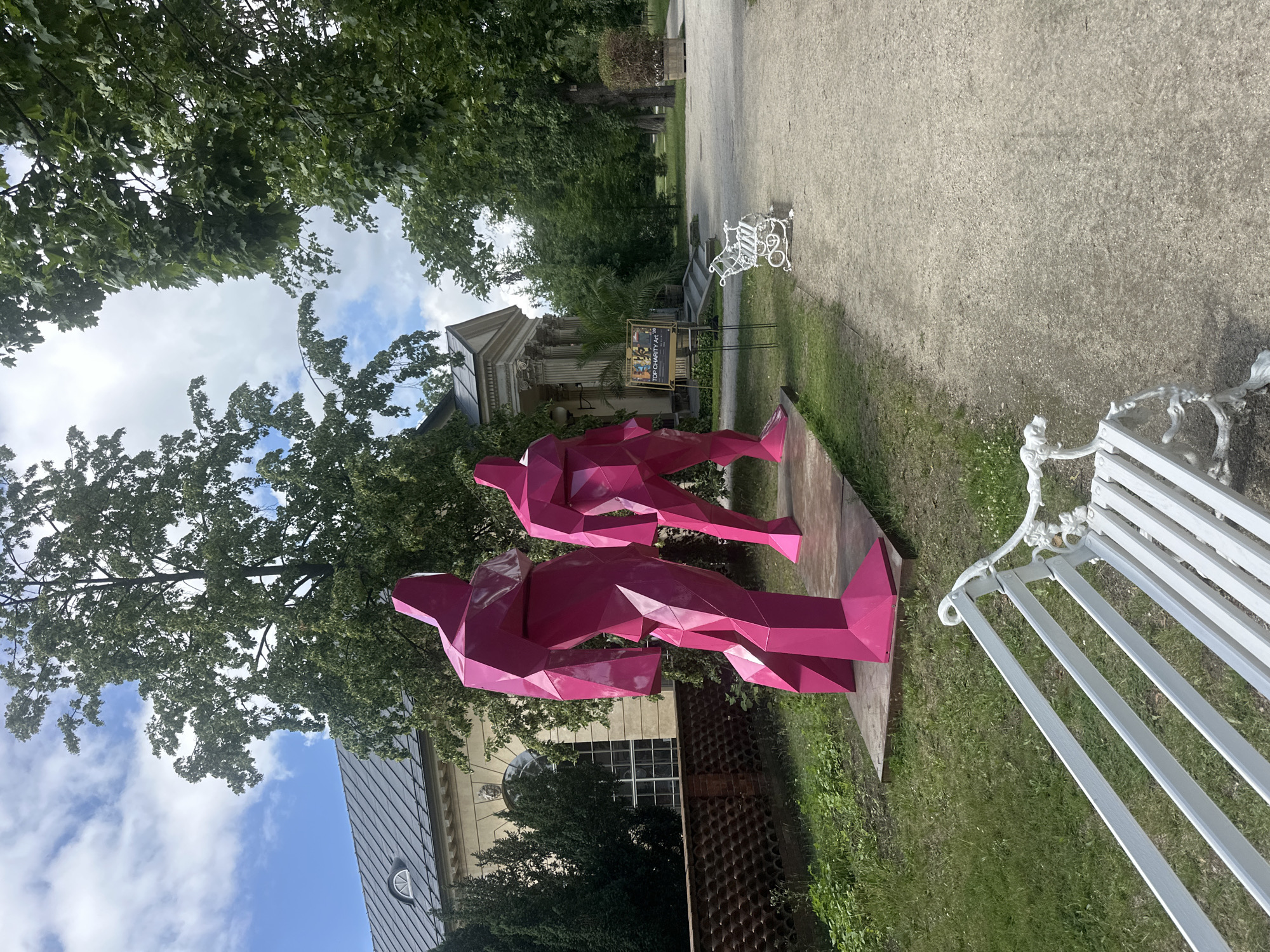 Two large, faceted, bright pink human-like sculptures stand in a grassy area next to a paved walkway in front of a building with a classical facade. The sculpture on the left is positioned to the left of the sculpture on the right, and both are facing forward. The building behind them has large windows and ornate architectural details. A white park bench is visible in the foreground to the left, and another white park bench is visible on the right side of the walkway further in the distance. Trees with green foliage are present throughout the scene. A sign that reads "TOP CHARITY Art 20" is visible between the sculptures and the building. This scene is located in Łazienki Park, Warsaw, Poland.