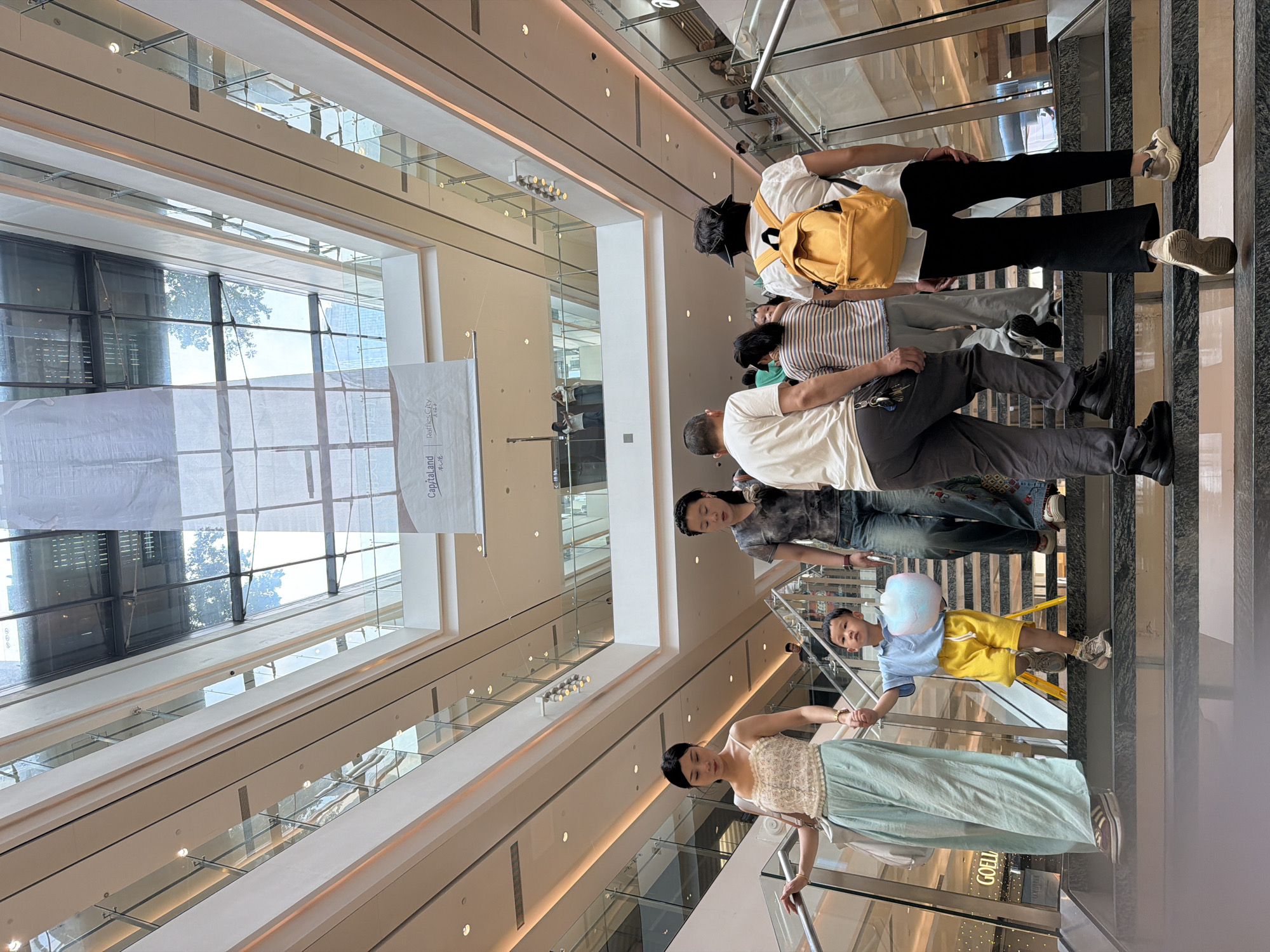 An upward angle shot shows people descending an escalator and stairs inside a multi-level modern shopping mall. The mall features a large, atrium-like central space with a glass ceiling, allowing natural light to filter in. The architecture is characterized by clean lines, white walls, and polished surfaces. Several levels are visible, with glass railings and escalators connecting them. A banner with Chinese text and a logo for "Capitaland Raffles City" is partially visible hanging in the upper part of the atrium.
In the foreground, a woman in a light floral top and a long, flowing light blue skirt is walking down the stairs, holding the hand of a young boy. The boy, wearing a light blue t-shirt and yellow shorts, is holding a large, fluffy pink cotton candy. Behind them, other shoppers are also descending the stairs and escalator. A woman in a grey tie-dye t-shirt and jeans is walking down the stairs to the right of the first woman. Further back, more people are visible on the stairs and escalators, some looking up towards the atrium. The atmosphere appears to be one of casual shopping and activity.