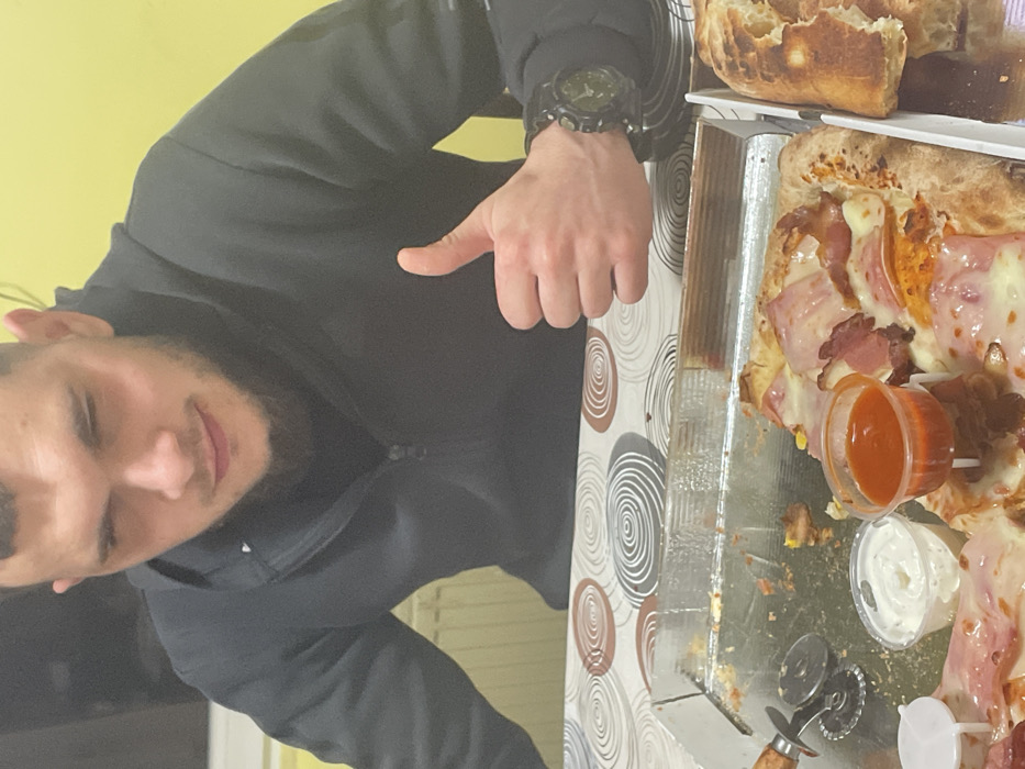 An indoor scene depicts a male subject seated at a table, stated to be located in Gorna Mitropolia, Bulgaria. The man, visible from the chest up, has short dark hair, a beard, and a mustache. He wears a dark, possibly black, hooded sweatshirt with a half-zip closure and a dark undershirt. A dark-colored rugged watch is on his left wrist. His left hand is raised in a 'thumbs up' gesture. His facial expression indicates a slight smirk or mild amusement, with his right eye slightly narrowed.
On the table, covered by a tablecloth with a brown and white concentric circle pattern, are multiple food items. A white rectangular pizza box contains a partially eaten square-cut pizza topped with cheese, bacon, and other visible ingredients like corn. Inside the pizza box, next to the pizza, are a metal pizza cutter with a wooden handle and two small plastic cups of sauce: one white and one red. To the immediate right of the pizza box, another white cardboard box holds a partially consumed piece of rustic-style bread. The background wall is solid yellow, with a white radiator visible on the left side of the frame. The lighting is consistent with indoor artificial sources.