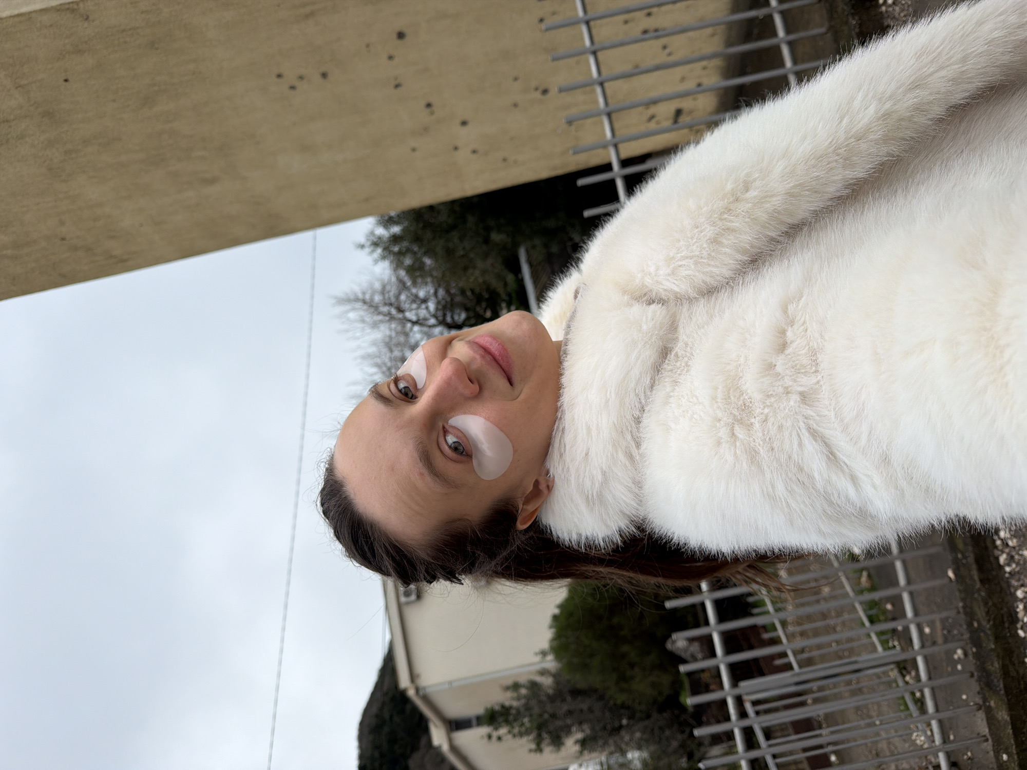 A female individual is captured outdoors in a medium shot, looking directly at the camera with a slight smile. She has dark brown hair, pulled back, and is wearing a thick, white faux fur coat. Two translucent, light-colored eye patches are visible on her lower eyelids.
The background on the left features a light-colored, multi-story building with a flat roof. Behind this building, a dark green, tree-covered mountain range is discernible under an overcast, light grey sky. Several bare deciduous trees are also present in the middle ground. On the right side of the frame, a large, tan-colored building or wall with visible surface imperfections occupies a significant portion of the background. A gray metal fence, consisting of vertical bars, runs along the bottom of the frame, separating the foreground from the mid-ground. The ground beneath the fence appears to be concrete or pavement, scattered with light-colored debris. The scene is located in the city of Budva, Montenegro.