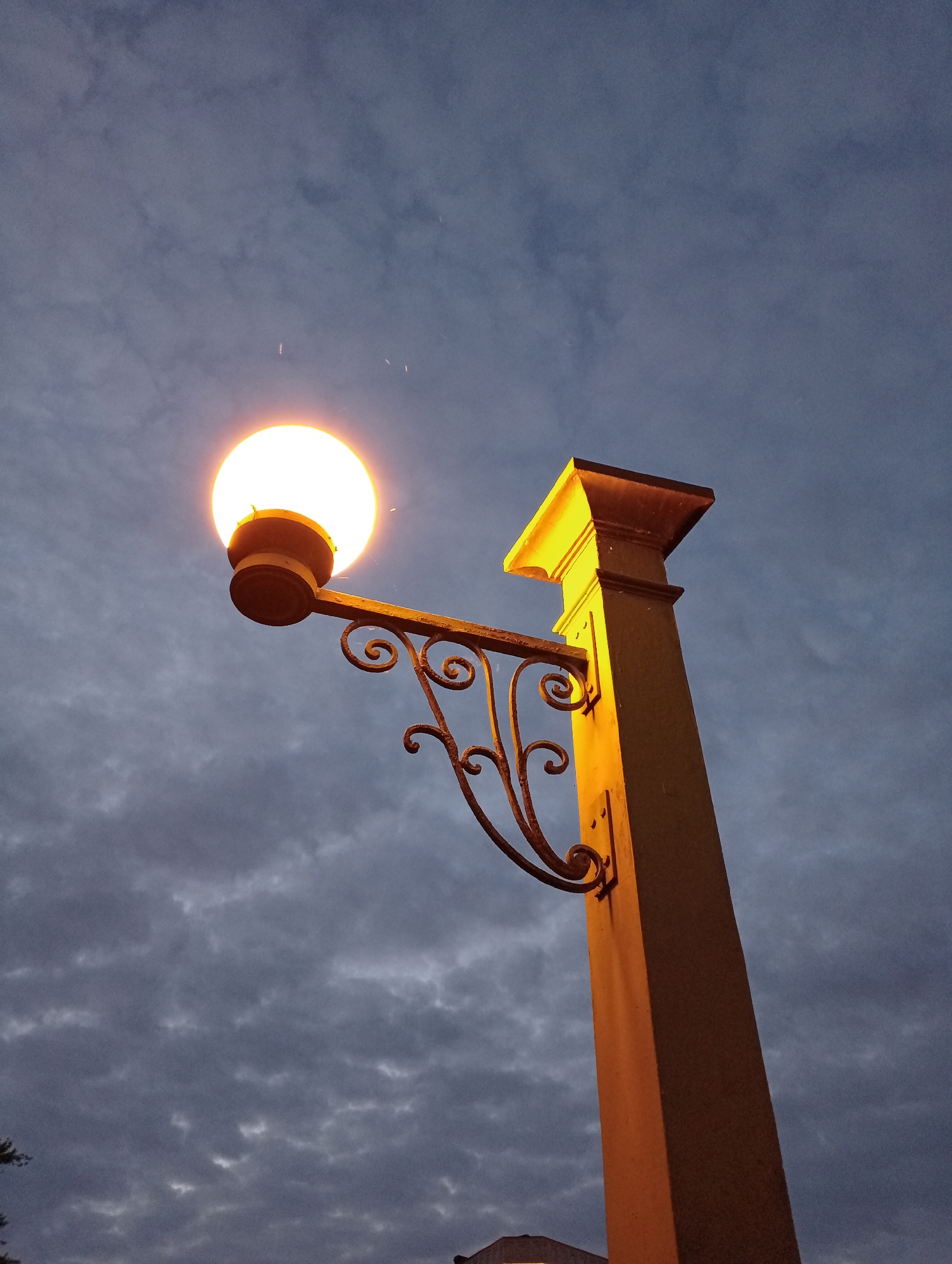 An outdoor, low-angle, medium shot captures a decorative lamppost against a dark, cloudy sky at dusk. The lamppost is made of a square, yellow-painted column topped with a pyramidal fixture. A black, wrought-iron arm extends from the column, supporting a round, illuminated lamp with a visible filament. The arm features ornate scrollwork. The sky is a deep blue with streaks of gray clouds, suggesting an overcast evening. There are no people visible in the image. The scene appears to be in a city setting, potentially along a street or in a park, though specific landmarks are not identifiable.