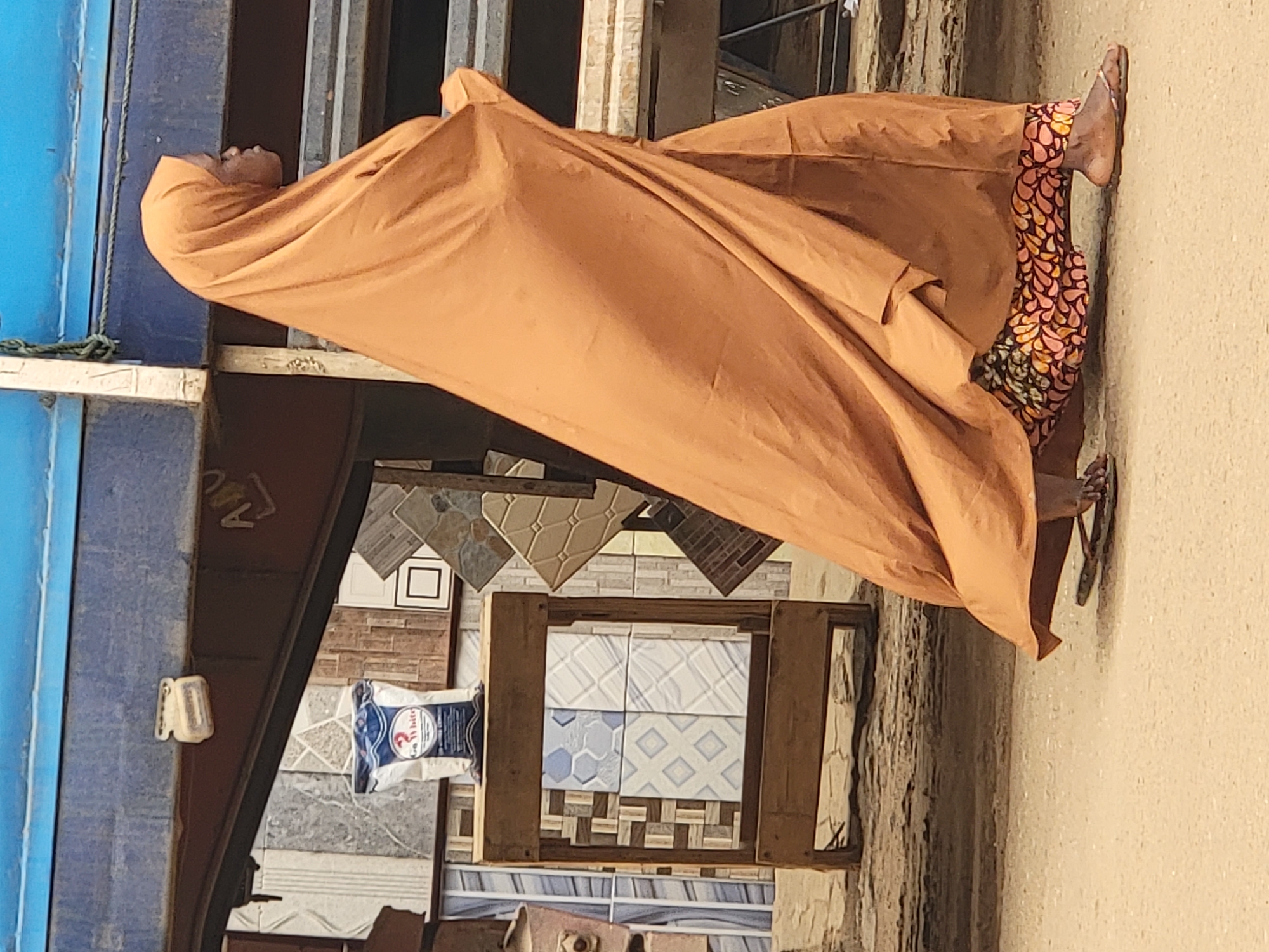 A person wearing an orange robe and patterned pants stands sideways near a building. The setting appears to be an outdoor area with a selection of decorative tiles and building materials in the background, suggesting a shop or market in the city of Maiburiji, Nigeria. The person's head is covered by the robe and they are wearing sandals on their feet.