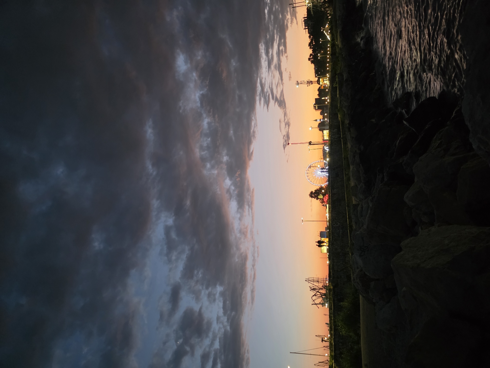 The image captures a sunset over the Varna Bay in Varna, Bulgaria. In the foreground, a rocky embankment meets the water, with waves gently lapping against the stones. The water is dark and reflects the fading light of the sky.
Dominating the middle ground is a seawall with a walkway, separating the water from the city's coastline. Silhouetted against the vibrant sunset sky are two figures walking side-by-side on the walkway. Beyond them, the Varna cityscape is visible, illuminated by numerous lights. Prominent structures include a Ferris wheel, several cranes, and other amusement park rides that are lit up. The sky above is a dramatic canvas of dark, heavy clouds tinged with the warm hues of orange, pink, and purple from the setting sun. A sliver of the moon is also visible in the upper left corner of the frame.