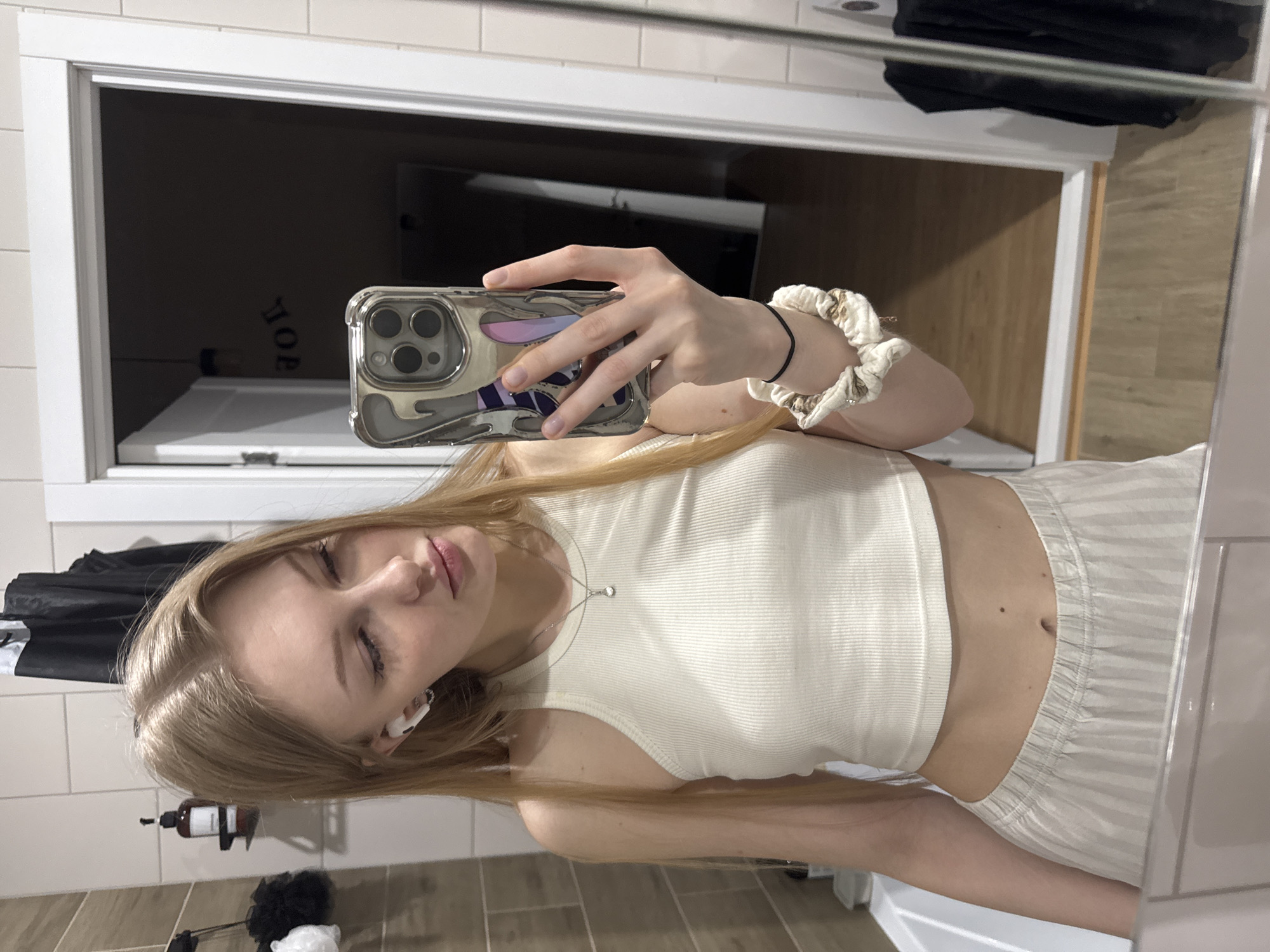 A young woman is taking a selfie in a bathroom. She is holding her phone in her right hand, with a scrunchie visible around her wrist. She is wearing a light-colored tank top and striped pants. The bathroom features tile walls and flooring, a vanity with a sink, and a mirror reflecting a doorway into another room. A dark object hangs in the reflection on the upper right of the mirror. There is a bottle of liquid soap and a mesh sponge. I am unable to confirm the location in Perm, Russia from this image alone.