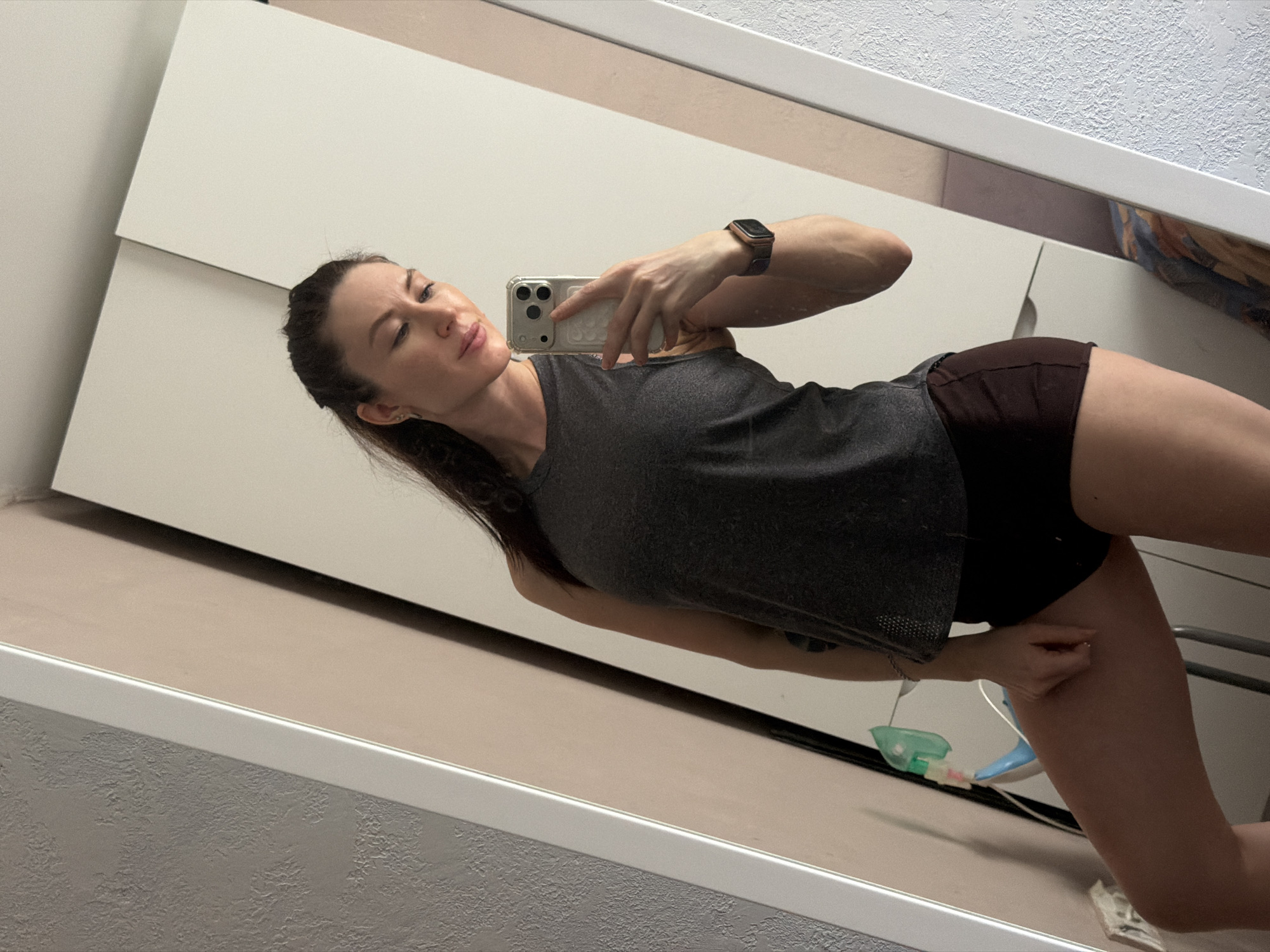 The image captures a mirror selfie of a woman in an indoor setting, likely in Perm, Russia. The shot is tilted, presenting the scene diagonally.
The central subject is a woman with long, dark hair, seemingly pulled back, and fair skin. She is dressed in a dark grey, possibly athletic, tank top and dark brown shorts. On her left wrist, she wears a smartwatch with a dark band. She holds a light-colored smartphone, equipped with multiple camera lenses and a pop-socket grip, in her right hand to take the photo. Her expression is neutral and calm, with her gaze directed slightly away from the camera. Her arms appear toned.
The background, seen through the mirror, reveals elements of a modern interior. Large, light-colored (off-white) wardrobe doors or cabinet panels are visible behind the woman. The lower section of the mirror reflects a textured wall, possibly stucco, painted in a light grey or beige, with a distinct white trim line. Below this, a light beige surface, likely the floor, is also reflected. Near her right hip, a small, multi-colored object with green and blue parts and some tubing is partially visible on the floor, but its specific function is not discernible.
The overall scene suggests a private, casual moment, possibly in a home environment like a bedroom or dressing room, captured without any visible text.