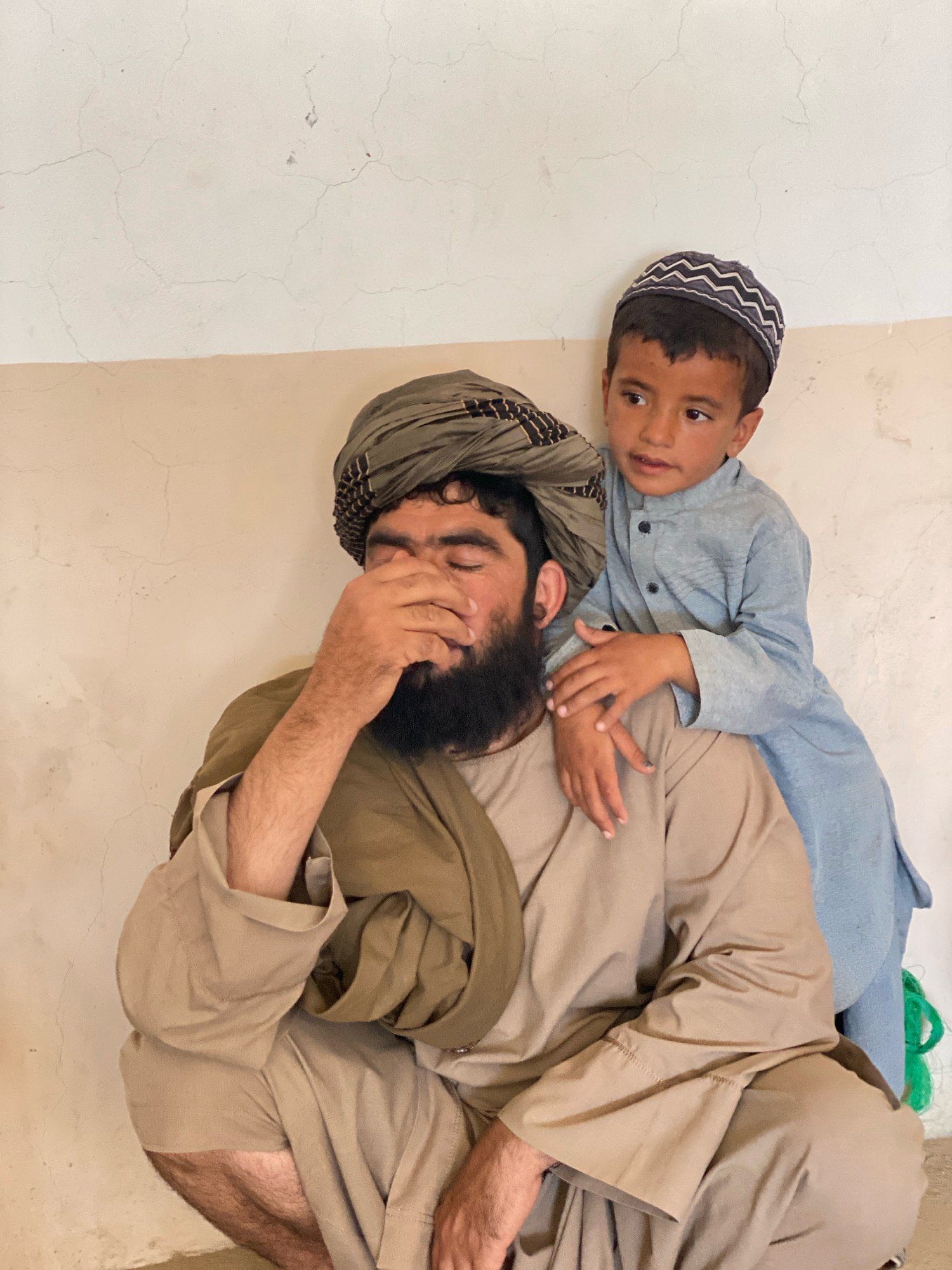 A low-angle, medium shot captures a man and a young boy in Pasab, Afghanistan. The man, crouched low, wears a tan garment and a large olive-green turban. His right hand covers his face, obscuring his eyes. His beard is dark and full. Behind him, the boy, wearing a light blue tunic and a patterned cap, stands with his arms around the man's shoulders, looking to his right. The background consists of a neutral-colored wall with visible cracks. A green object is partially visible on the right side of the frame. The lighting is bright and even.