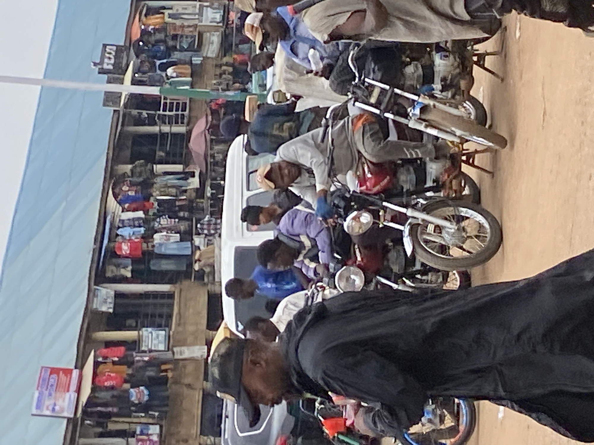 This image captures a bustling street scene in what appears to be a market or commercial area in Bukuru, Nigeria. The photograph, oriented correctly, shows a busy thoroughfare lined with multi-story buildings that serve as shops or stalls, displaying various goods such as clothing and packaged items.
Numerous people, predominantly men, are present throughout the scene. They are casually dressed in t-shirts, shirts, and trousers, with some wearing caps or hats. Many individuals are either sitting on or standing next to motorcycles, suggesting the operation of motorcycle taxi services (known as 'okada') or simply a common mode of transport. A white van or mini-bus is also visible, surrounded by people, indicating a general gathering point or transport hub.
The ground appears to be unpaved, dusty earth, consistent with such local street environments. The lighting suggests it is daytime, though the exact time is not clear. The overall atmosphere is active and dynamic, reflecting the daily life of a busy market street.
A notable detail is a sign on a building in the upper left portion of the image, where the word "FUDOS" can be discerned. Other signs and texts are too blurry to read clearly. No strong emotions are visible on the faces of the people, who appear to be engaged in their daily activities.