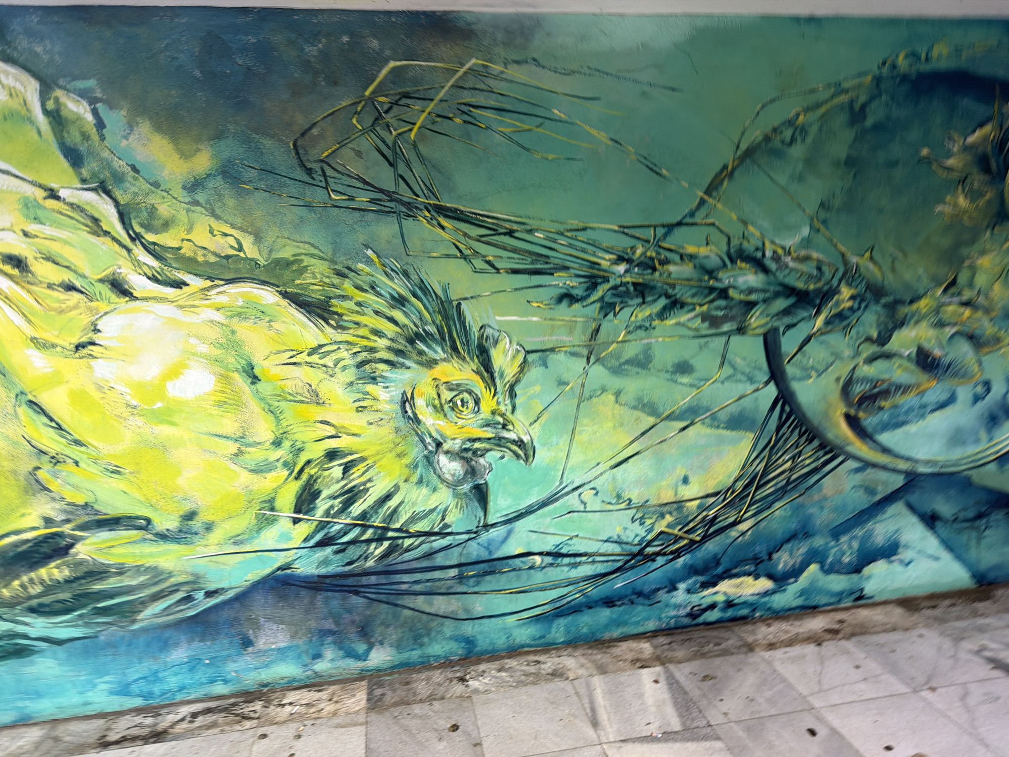 The image depicts a mural, likely located in Sofia, Bulgaria. The artwork features a stylized yellow and green rooster as the central subject. The background incorporates swirling lines and abstract shapes in shades of green and blue. Some linear elements resemble wires or branches, adding complexity to the composition. The mural is painted on a wall adjacent to a tiled sidewalk. The photograph is taken at an angle, suggesting the mural is on a wall near ground level. The overall scene conveys an urban art installation with a focus on nature and abstract design elements.