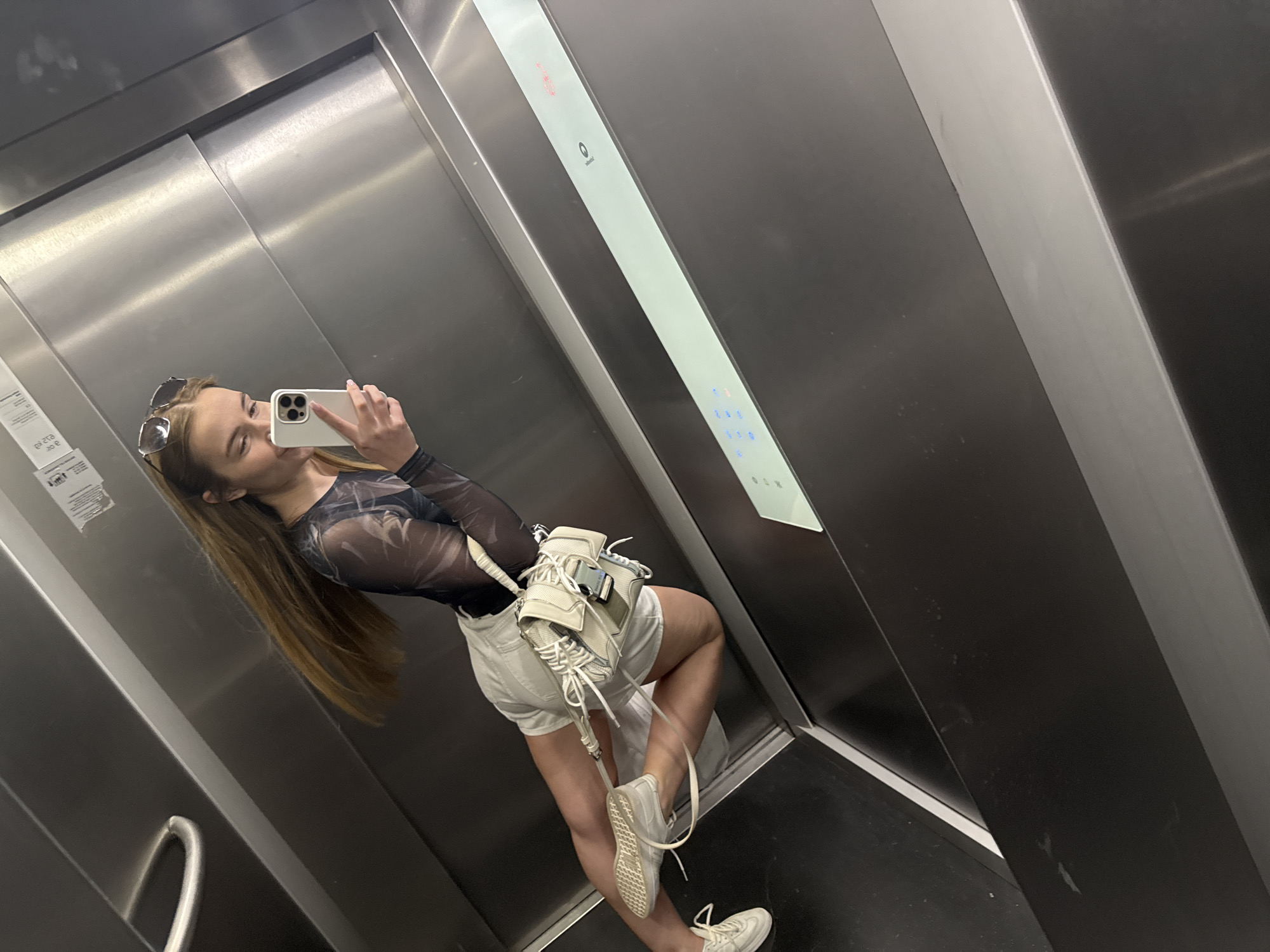 A young woman is taking a selfie in an elevator. She is wearing a mesh top, white shorts, and white sneakers, and has a white bag over her shoulder. She is holding her phone in her right hand, angled towards the mirror surface of the elevator. The elevator has silver walls and black floors. There's a control panel to the right, with illuminated buttons. Based on the architectural style and available visual information, the location is possibly in a building in Budva, Montenegro, however, further evidence is needed to confirm this.