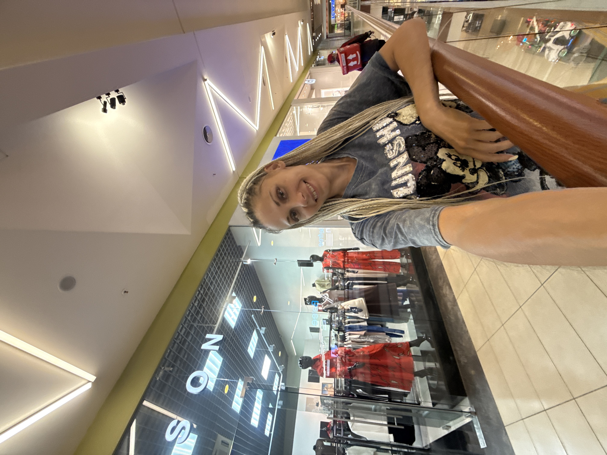 A young woman with blonde braided hair smiles for a selfie in a brightly lit shopping mall. She is wearing a gray t-shirt with a graphic design that includes the word "SUNSHINE" in sparkling letters and a sequined Mickey Mouse. The woman is leaning on a wooden railing, which runs along a balcony or upper level of the mall. Below her, mannequins in a store window display red and white clothing. The mall's architecture features modern geometric designs with white panels and integrated lighting. In the background, more shops and escalators are visible, suggesting a bustling commercial environment. The lighting suggests it is daytime, and the indoor setting implies controlled weather conditions. A Russian flag is not visible, but the location is identified as Arkhangelsk, Russia.