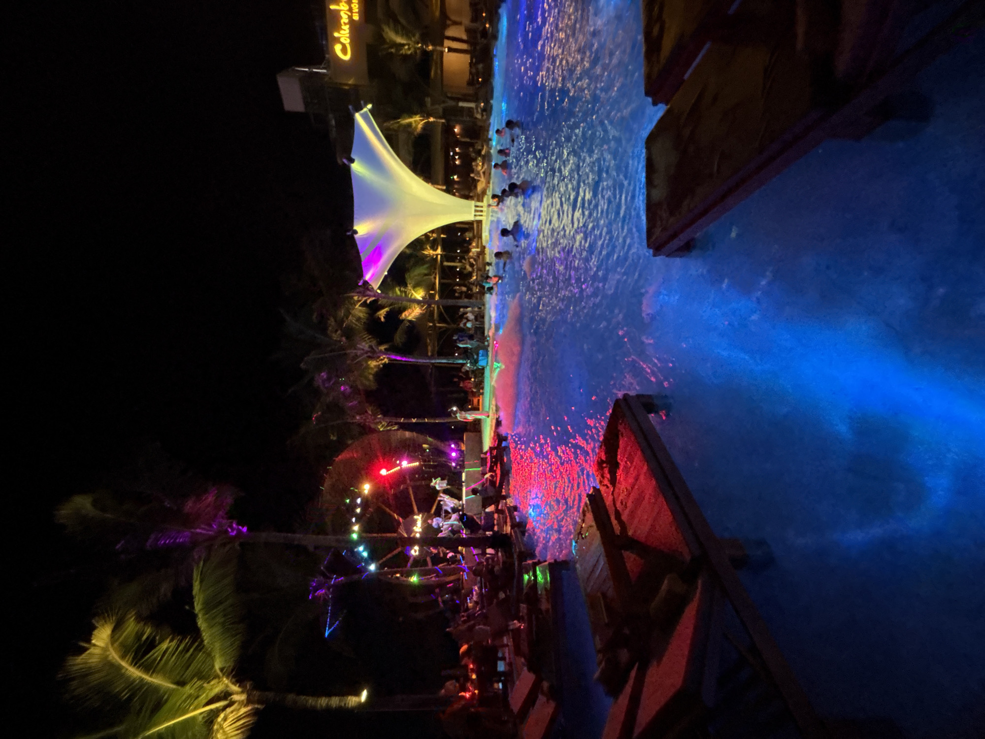 The image depicts a vibrant nighttime scene at a resort swimming pool, likely in Đà Nẵng, Vietnam, given the presence of palm trees and a tropical atmosphere. In the foreground, red-lit lounge chairs are partially submerged in the pool, casting a warm glow on the blue water. The pool itself is illuminated with blue and pink lights, creating a dynamic and festive ambiance.
In the mid-ground, a group of people are enjoying themselves in the water, some appearing to be dancing or socializing. Further back, a stage area is set up with performers on it, illuminated by colorful stage lights, suggesting a live music or entertainment event. Palm trees with illuminated fronds frame the scene on the left and right, adding to the tropical setting. In the background, a building with a prominent yellow sign displaying "Columb..." is visible, indicating a resort or establishment. The overall atmosphere is lively and suggests a popular nightlife or entertainment venue.