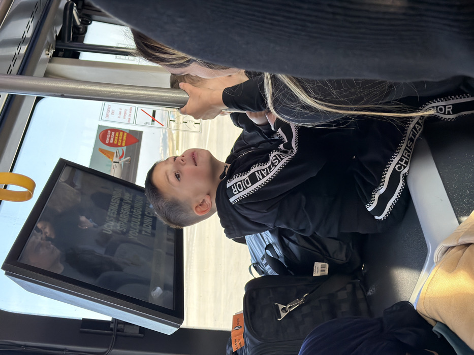 The scene depicts the interior of an airport shuttle bus, positioned on an airfield tarmac at Sabiha Gökçen International Airport (SAW) in Pendik, Istanbul, Turkey, a major transportation hub for the adjacent district of Tuzla.
In the foreground, a young male child, approximately 4-6 years old, is seated on a dark surface, possibly a bench or luggage area, looking upwards and slightly to the right with a soft, open-mouthed expression. He has short, dark hair and is wearing a black tracksuit featuring a white and black striped pattern with "CHRISTIAN DIOR" lettering along the sleeves and lower pant legs. To the child's left, multiple pieces of dark luggage are stacked, including a black checkered rolling suitcase with an orange "CABIN & MAIN BAGGAGE" tag, and a black duffel bag.
On the right side of the frame, partially obscured, is an adult individual, likely female, whose blonde hair and a hand gripping a vertical metal support pole are visible. This suggests the adult is either standing or steadying themselves within the moving vehicle. Reflections of other passengers are faintly visible on the window and an overhead screen.
An angularly mounted rectangular display screen is positioned above the child, reflecting interior elements and displaying Turkish text that translates to "Order during flight, enjoy your journey." Adjacent to the screen, visible through the window, are several informational decals: a red, heart-shaped advertisement featuring a white airplane and "PEGASUS" branding, an "EMERGENCY EXIT" sign in English with instructions to "break the window," and a standard 'no smoking' symbol.
Outside the window, a brightly lit airfield tarmac is visible under a clear sky. Distant airport infrastructure and what appears to be the tail or wing of an aircraft can be discerned, consistent with an active airport environment. Yellow grab handles are visible along the bus ceiling.
