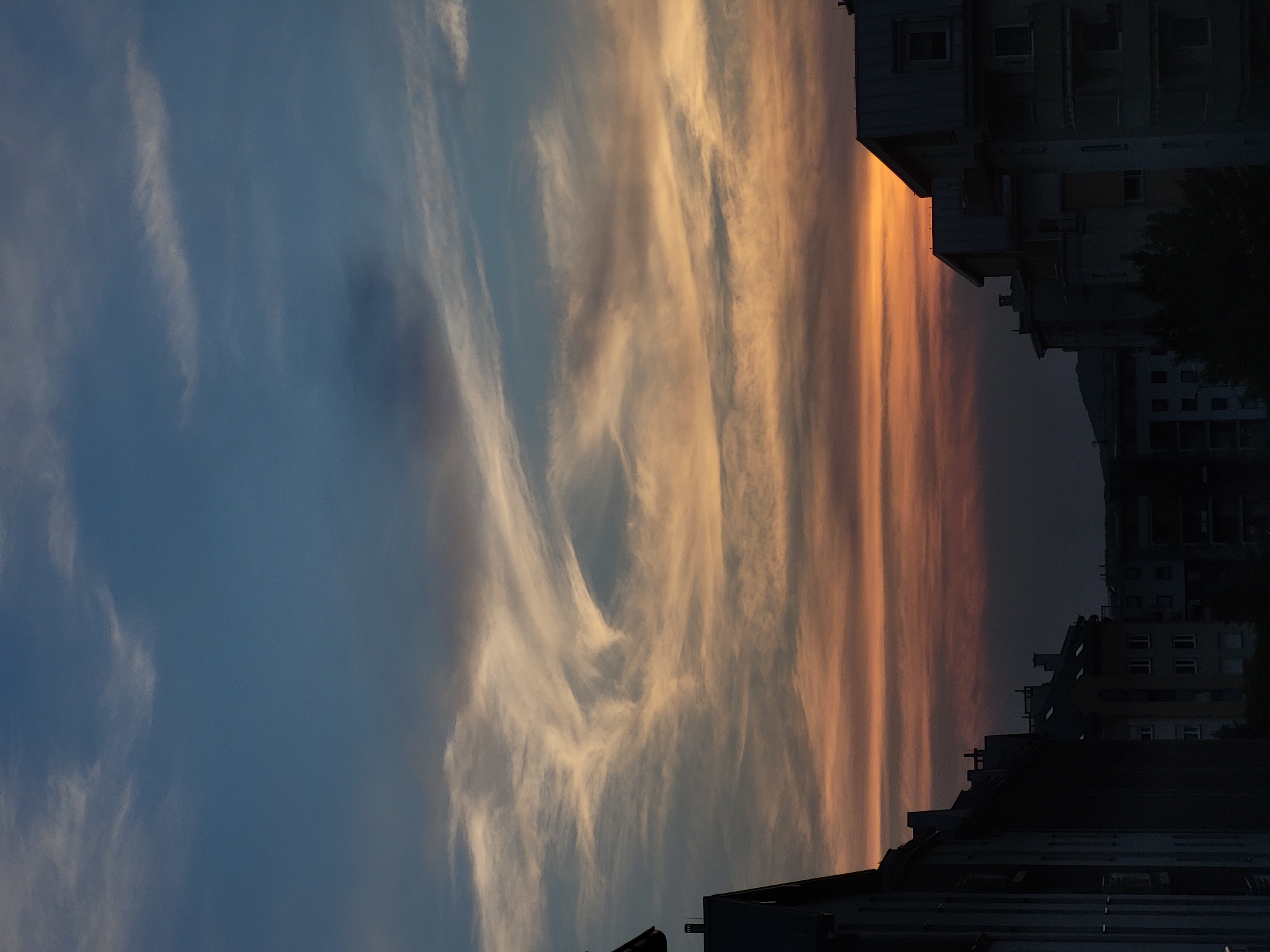 The image depicts a twilight sky over an urban landscape. In the foreground, the silhouettes of several buildings are visible, their dark forms contrasting with the illuminated sky. The sky itself is a gradient of colors, transitioning from a deep blue at the top to bands of orange and red near the horizon, indicative of sunset or sunrise. Wispy clouds stretch across the sky, catching the light and creating intricate patterns. The buildings appear to be multi-story structures, some with visible rooflines and windows, suggesting an apartment or residential area. The scene is devoid of people or any discernible activity, presenting a quiet, atmospheric view of a city at dusk.