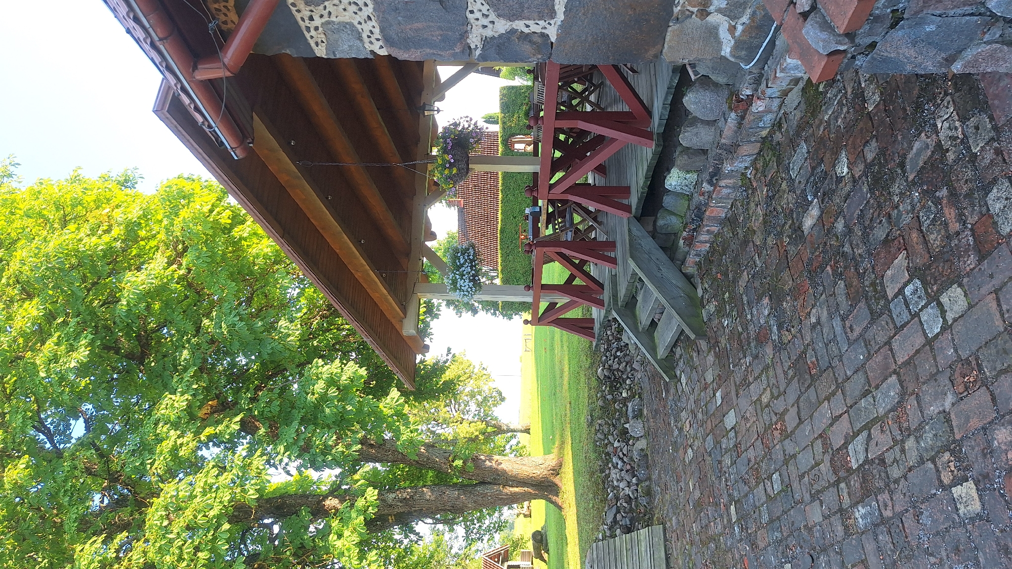 The image shows a covered outdoor seating area with wooden tables and chairs under a pitched wooden roof supported by posts. Two hanging baskets filled with flowers are suspended from the roof. The area is accessed by wooden steps and is bordered by a red wooden railing. The flooring is made of bricks. To the left, a large tree with lush green leaves stands prominently, and a wooden pathway leads away from the seating area. In the background, a manicured hedge and a red-tiled roof of a building are visible, suggesting a rural or semi-rural setting. The sky is clear and bright, indicating daytime. This scene is likely located in the Lēdmane municipality, Latvia, given the presence of traditional Latvian countryside elements.