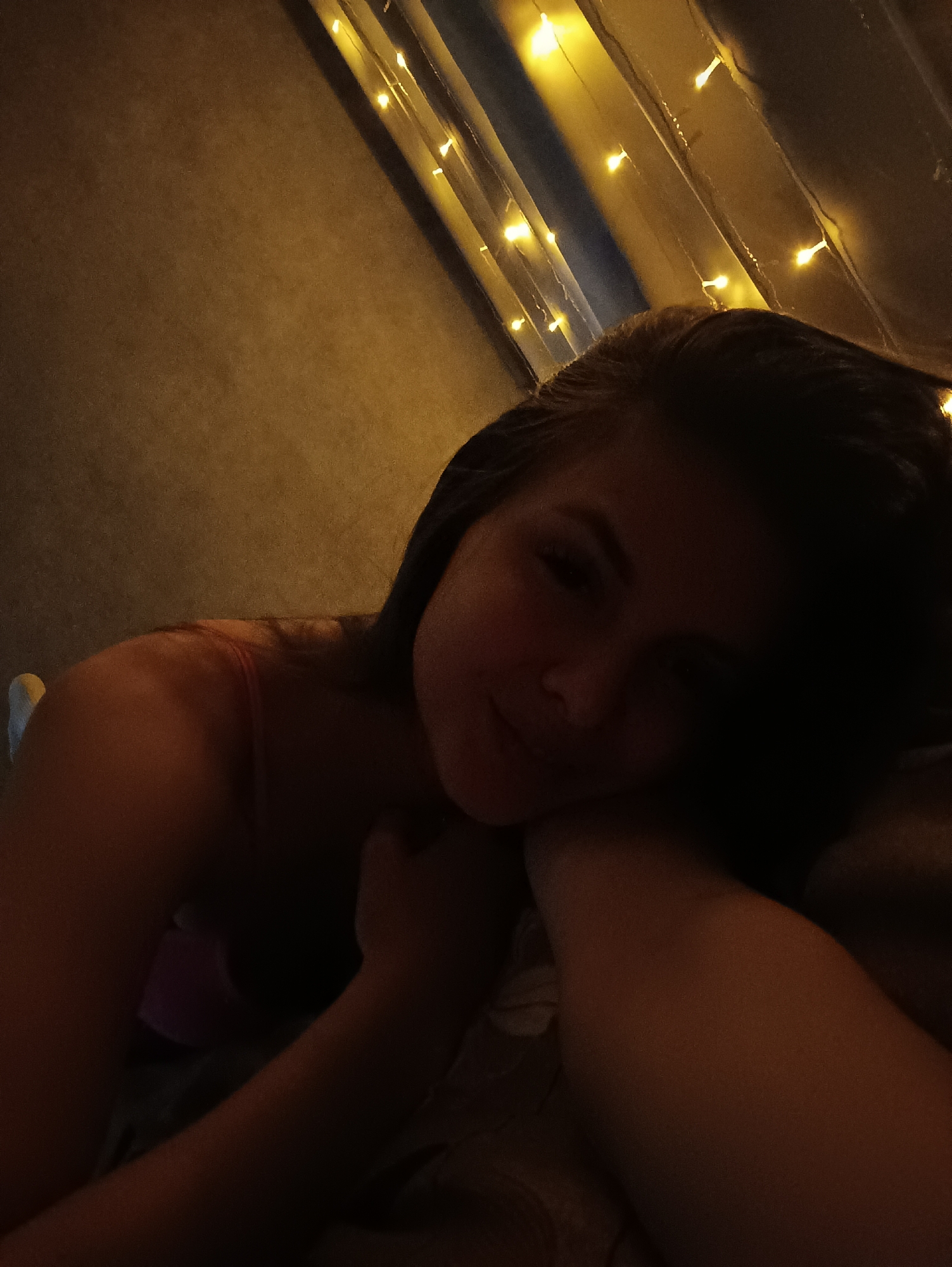 A woman with long dark hair is resting her head on her arm, possibly in a bed or on a soft surface. The lighting is dim, primarily illuminated by string lights in the background. The background appears to be a wall, possibly draped with fabric. There is no immediate indication of the location being Chaikovsky, Russia within the image itself. No other persons, objects, or actions can be determined from the image.
