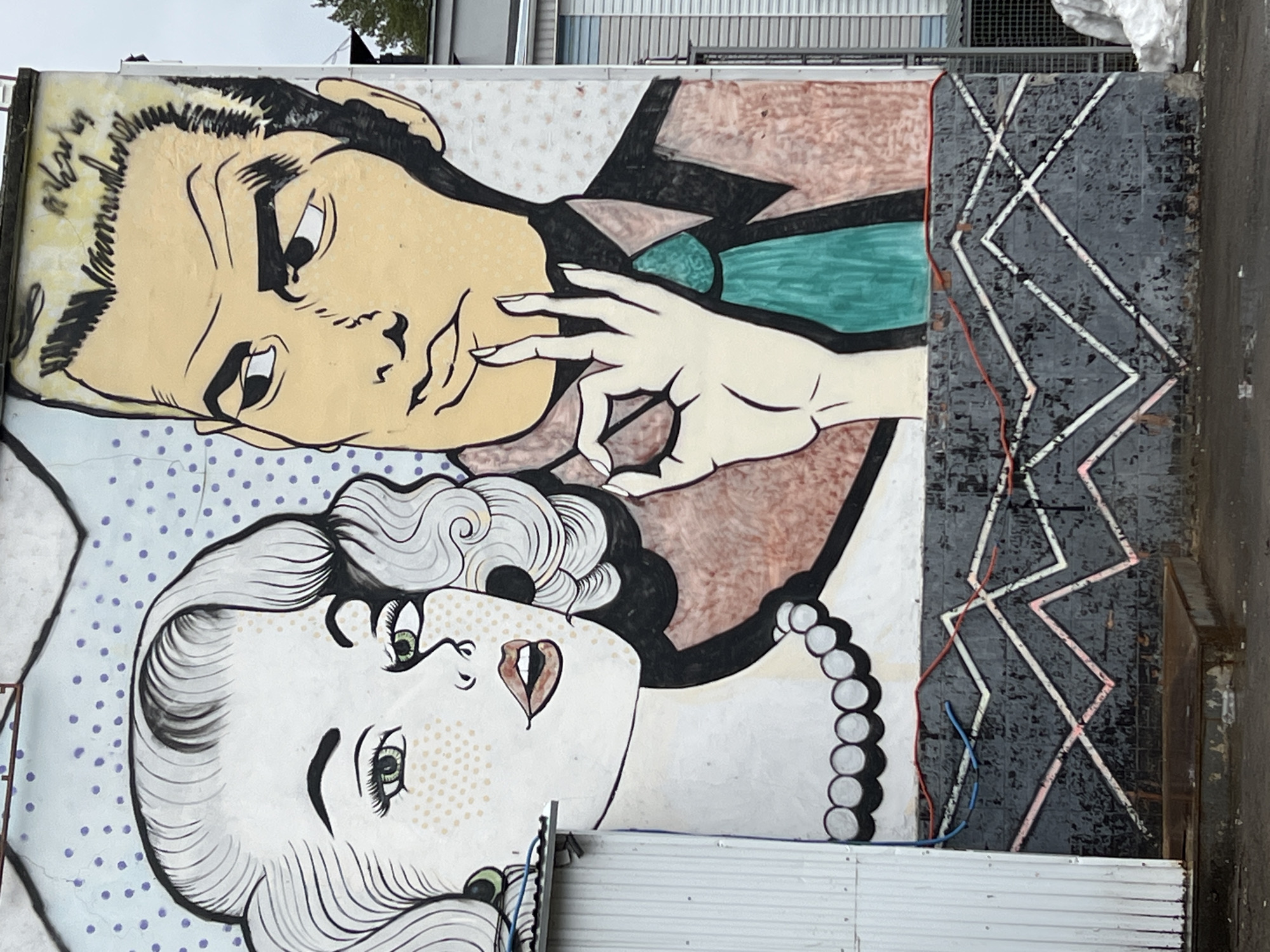 A large mural is affixed to the side of a building. The mural depicts two figures in a style reminiscent of pop art. The first figure is a man with blond hair, eyes cast downward. The second figure is a woman, positioned below the man. She has white hair with dark accents, and her face features stylized freckles and makeup. The mural extends to the edge of the building and then transitions to a geometric black, white, and gray pattern. The bottom of the mural meets a white and corrugated surface. The location of this mural is in the city of Chaikovsky, Russia.
