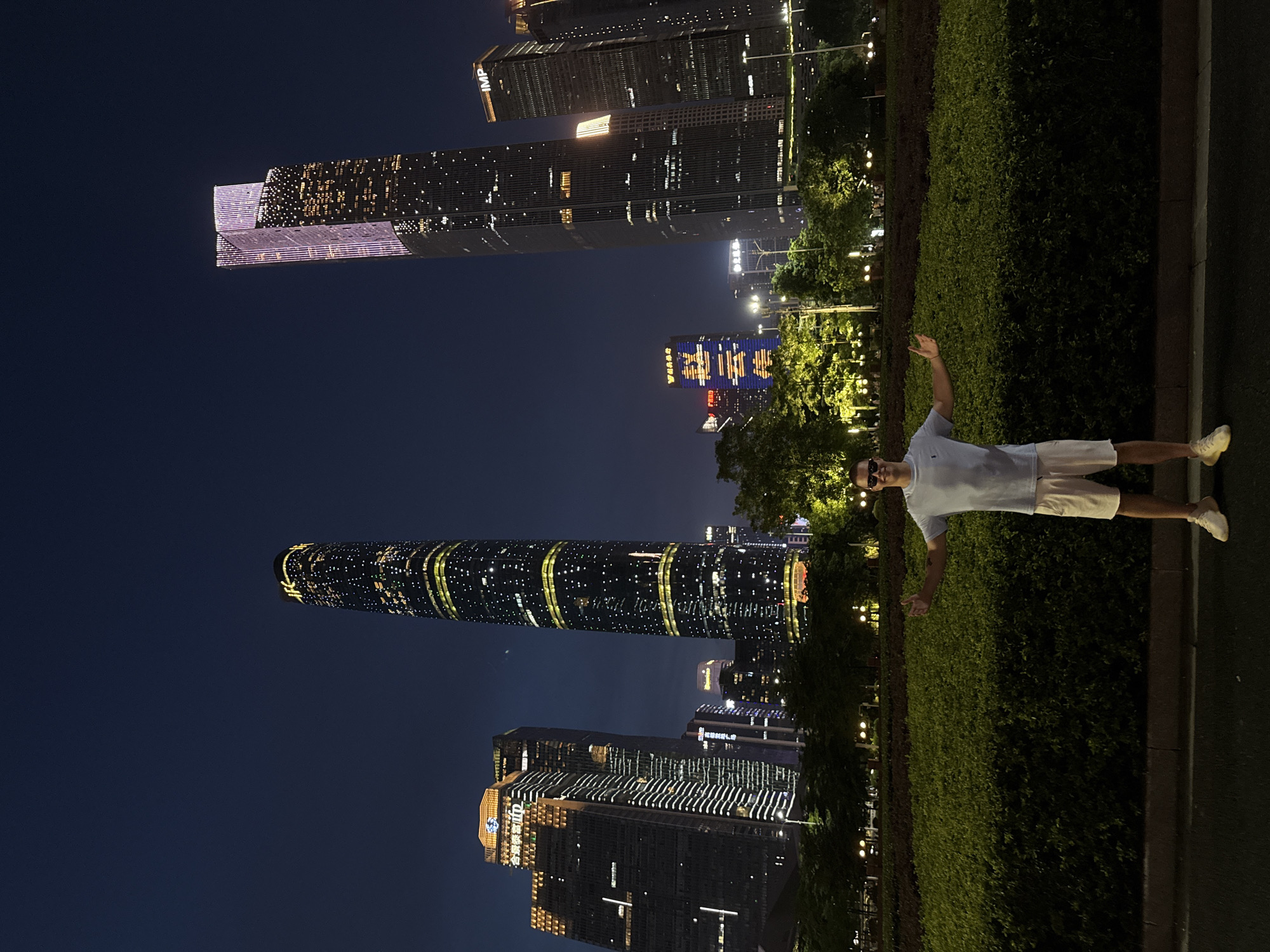 The image depicts a nighttime scene in Tianhe District, China, with a person standing in the foreground and illuminated skyscrapers in the background. The person, a male, is wearing a light blue polo shirt and white shorts, with white sneakers. He has his arms outstretched to his sides, palms up, and is looking towards the camera with a smile. He is positioned in front of a manicured green hedge, which is the dominant feature of the mid-ground.
The background is dominated by a cluster of modern skyscrapers, brightly lit and reflecting the night sky. Notable buildings include a very tall, slender skyscraper with a distinctive pattern of yellow lights and horizontal bands, and another taller skyscraper with a dual-peaked roof, illuminated with purple and white lights. Other buildings, some with illuminated signage, are also visible. Trees and streetlights are interspersed between the person and the buildings, adding depth to the scene. The overall atmosphere is one of urban nighttime illumination and a tourist posing for a photo.
