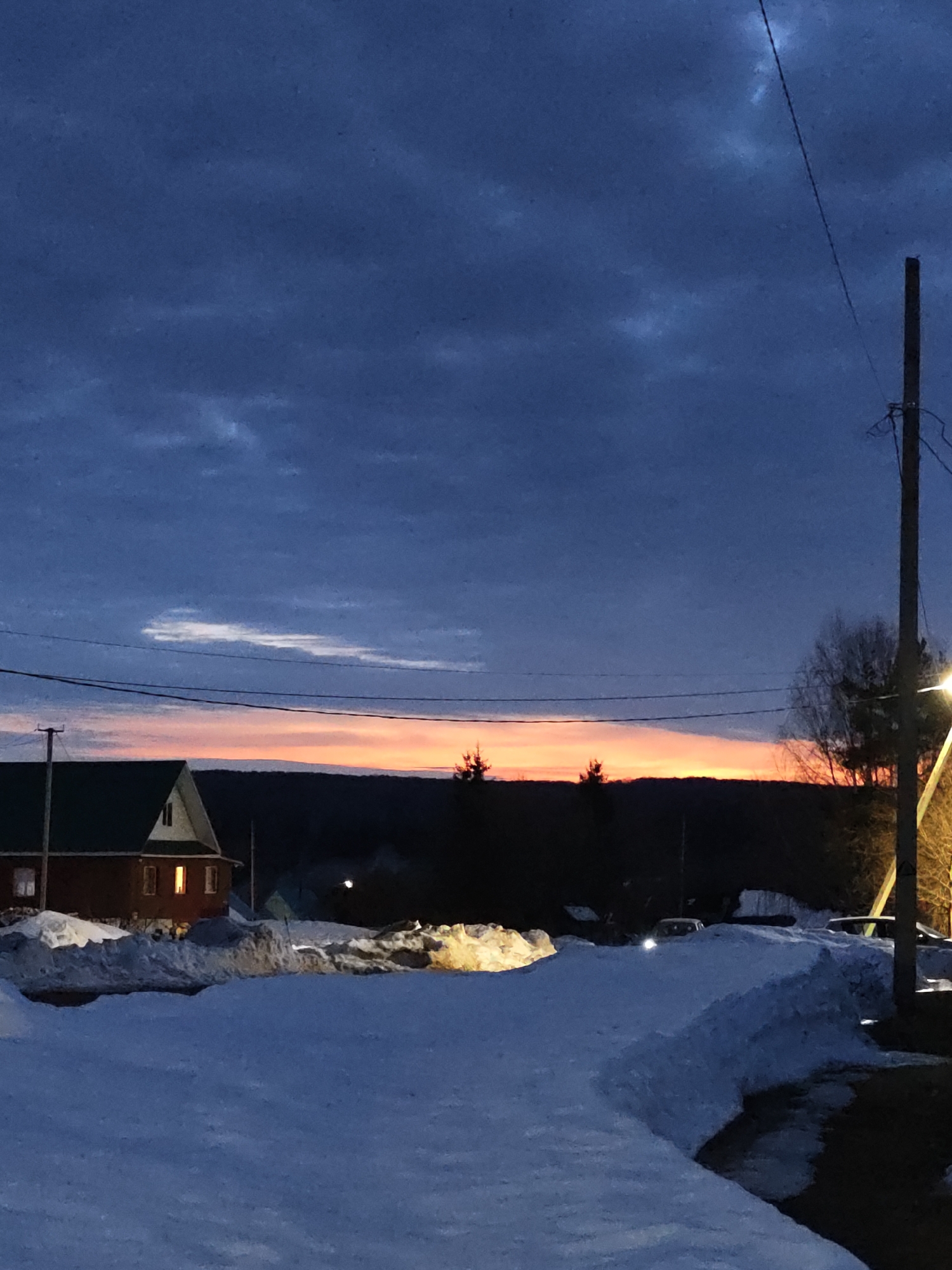 The image shows a winter scene in Уральское, Russia at dusk. A snow-covered road is visible in the foreground, flanked by large piles of snow on either side. A small, brick house with a green roof and lit windows sits to the left of the road. In the distance, a dark silhouette of trees and a hill is visible against a narrow strip of orange-pink sunset. A utility pole stands on the right side of the frame with power lines stretching across the sky. The sky is predominantly a dark blue-grey with textured clouds. There are vehicles parked behind the mounds of snow.