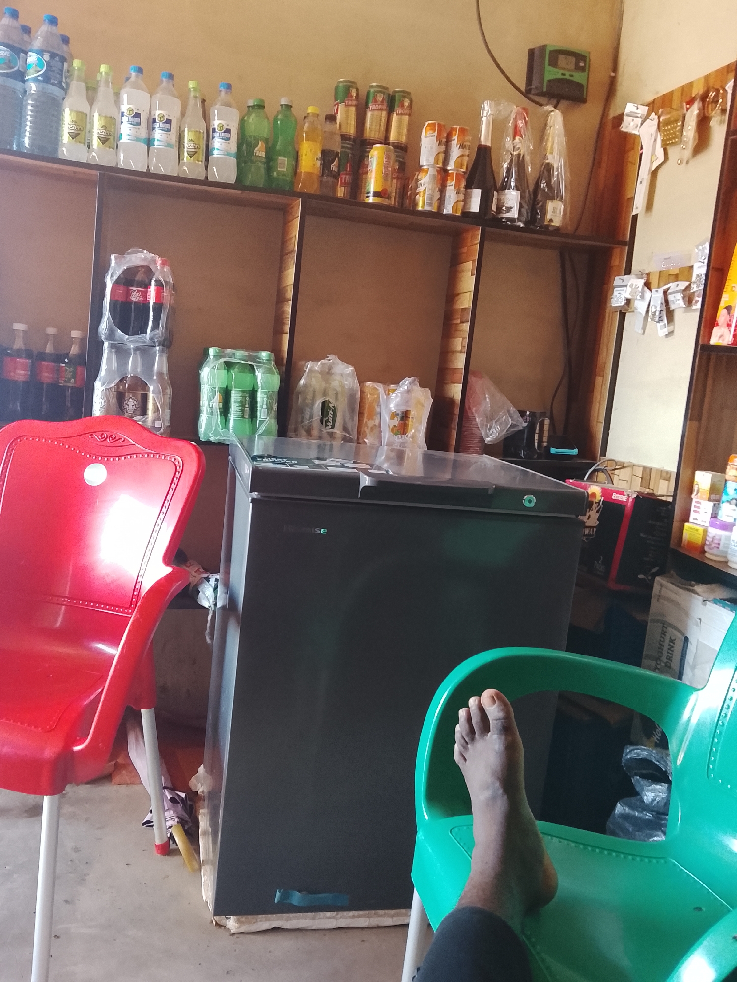 This media file depicts an indoor scene within a small shop or convenience store, likely located in Durumi, Nigeria. The setting is characterized by shelves stocked with a variety of beverages, including bottled water, soft drinks, and what appear to be canned alcoholic beverages. Several bottles of what seem to be sparkling wine or champagne are also visible on the upper shelf, some individually wrapped in plastic.
In the foreground, two plastic chairs are prominent: one is a bright red chair to the left, and a vibrant green chair to the right. A person's bare foot is resting on the seat of the green chair, suggesting someone is seated there, though the rest of their body is not visible. Behind the chairs, a dark-colored refrigerator or freezer unit is partially visible.
On the shelves, various products are displayed, with some branding visible. For example, there are stacks of green-bottled beverages, and a few bottles of Coca-Cola are visible. Some packaged drinks, possibly juices or water, are also present. To the right of the shelves, a section of a wall is adorned with small items, possibly accessories or small household goods, attached to a board. A small green electronic device with a display is mounted on the wall above the shelves.
The lighting suggests it is daytime, with artificial light supplementing natural light, creating some reflections on the plastic items. The overall impression is of a modest retail establishment stocked for sale. There are no obvious cues about the weather outside or a specific event taking place, other than the everyday operation of a shop.