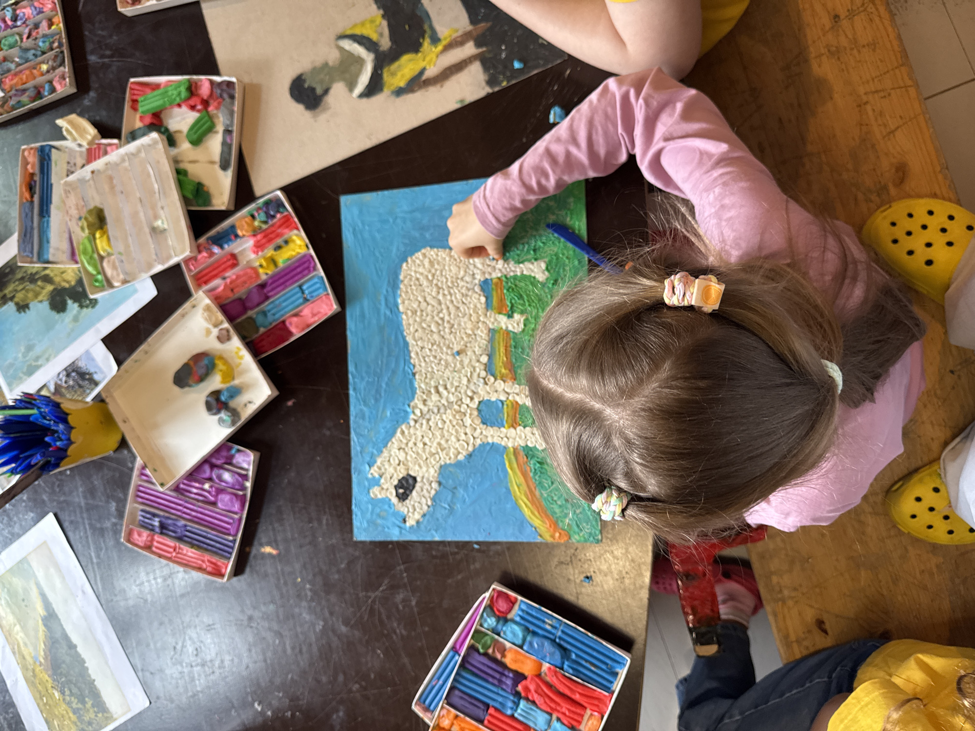 The overhead shot captures a close-up of a young girl in a pink long-sleeved shirt, her back to the camera, as she works on an art project at a wooden table. Her brown hair is styled in pigtails secured with colorful bands. In front of her, a rectangular artwork depicts a white sheep with a black face against a bright blue sky, green grass, and a rainbow. The sheep is textured with many small, light-colored pieces, possibly clay or rice grains. To the left of the artwork is an open box filled with colorful sticks of clay in shades of blue, purple, pink, and red.
Scattered on the table are various art supplies, including other open boxes containing clay in different colors, a yellow cup filled with blue and black markers or pens, and several framed pictures or art pieces. One artwork to the upper left features a landscape with muted colors. To the right, another piece of art shows a silhouetted figure in black and yellow.
The table surface appears to be a dark, textured wood. The girl's feet are visible at the bottom of the frame, wearing yellow Crocs with black polka dots. Another person's legs and feet, also wearing yellow Crocs, are visible on the left side of the image, suggesting a shared creative space. The overall scene depicts a moment of artistic activity, likely in a home, classroom, or art studio setting. The specific location in Moscow, Russia, cannot be determined from the visual information provided.