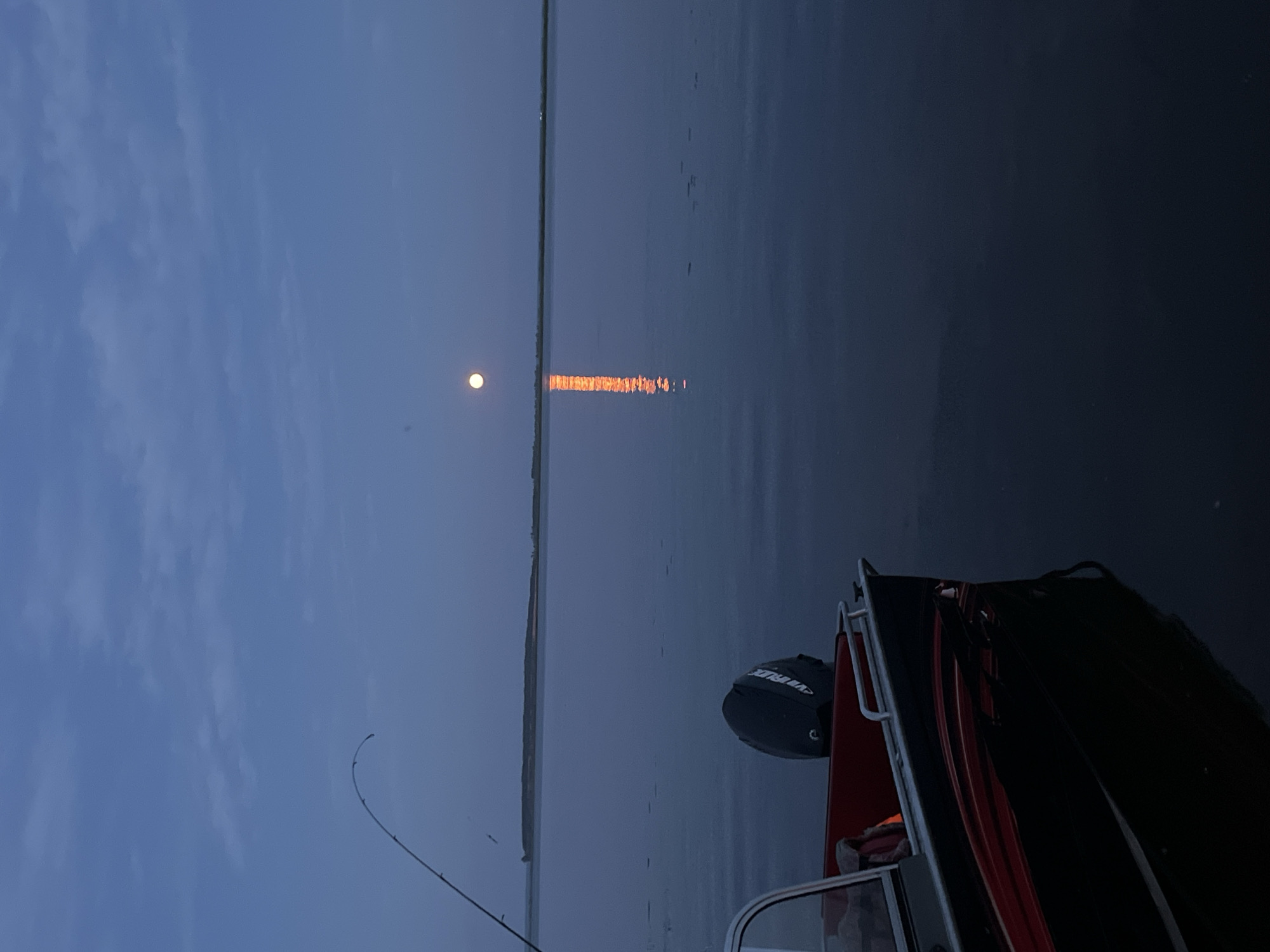 The image depicts a serene twilight scene on a body of water, possibly a river or lake, with a full moon visible in the dusky sky. A fishing rod extends from the left foreground, its tip arcing gently upwards. In the lower left corner, the bow of a dark-hulled boat with red accents is visible, featuring a black outboard motor at its stern. The moon casts a shimmering reflection on the calm water, creating a bright, elongated path of light. A distant, low-lying landmass is visible on the horizon. The sky is a deep, muted blue, with subtle cloud formations. The overall atmosphere is peaceful and tranquil. The location is identified as Ust-Belsk, Russia.