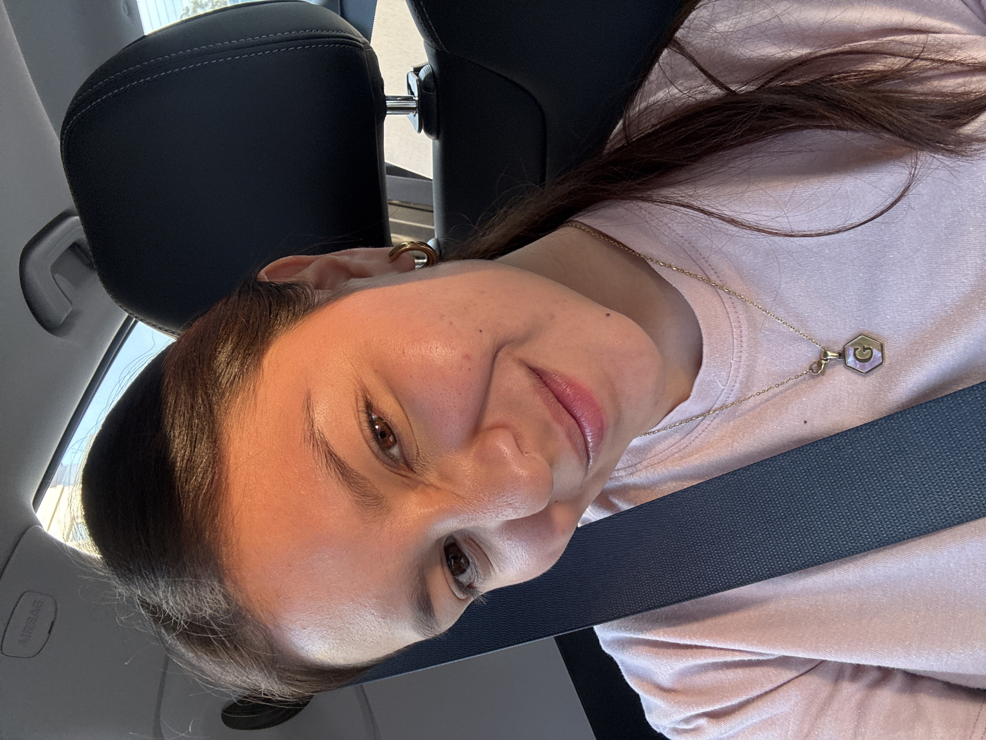 A woman with brown hair and brown eyes is in a car. She is wearing a seatbelt, a light pink shirt, and a gold necklace with a hexagonal pendant. She has a gold earring. The interior of the car is visible, including a black leather headrest. The photograph appears to be taken in daylight. There is no indication in the image that places it in the city of Puebla, Mexico.