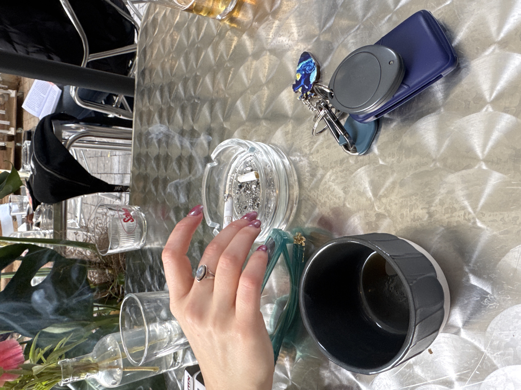 The scene depicts a metal-textured outdoor cafe table in Tel-Aviv, Israel, specifically identified by a "Salud" branded glass, consistent with Salud Cafe on Allenby Street.
In the foreground, a dark grey and white ceramic cup, containing a small amount of dark liquid, is positioned on the left. Adjacent to it, a pair of teal-colored eyeglasses rests on the table. A female's left hand, with pink-purple manicured nails and a silver ring featuring an oval-shaped stone on the middle finger, is centrally placed, hovering over a clear glass ashtray. The ashtray contains multiple cigarette butts and ash, with smoke visibly rising from a burning cigarette. To the right, a set of keys includes a blue remote fob, a grey oval fob, and a distinctive blue and yellow keychain depicting an stylized creature. A partial view of a glass with an amber liquid is visible on the far right.
In the background, behind the table, a clear glass bottle with pink flowers and green stems stands next to an empty clear glass. Large green foliage partially obstructs the view. Silver metal chairs are visible, one with a black bag draped over it. A person is seated, obscured partially by a chair, holding an open book. The background includes a light-colored paved area, indicative of a street or sidewalk.