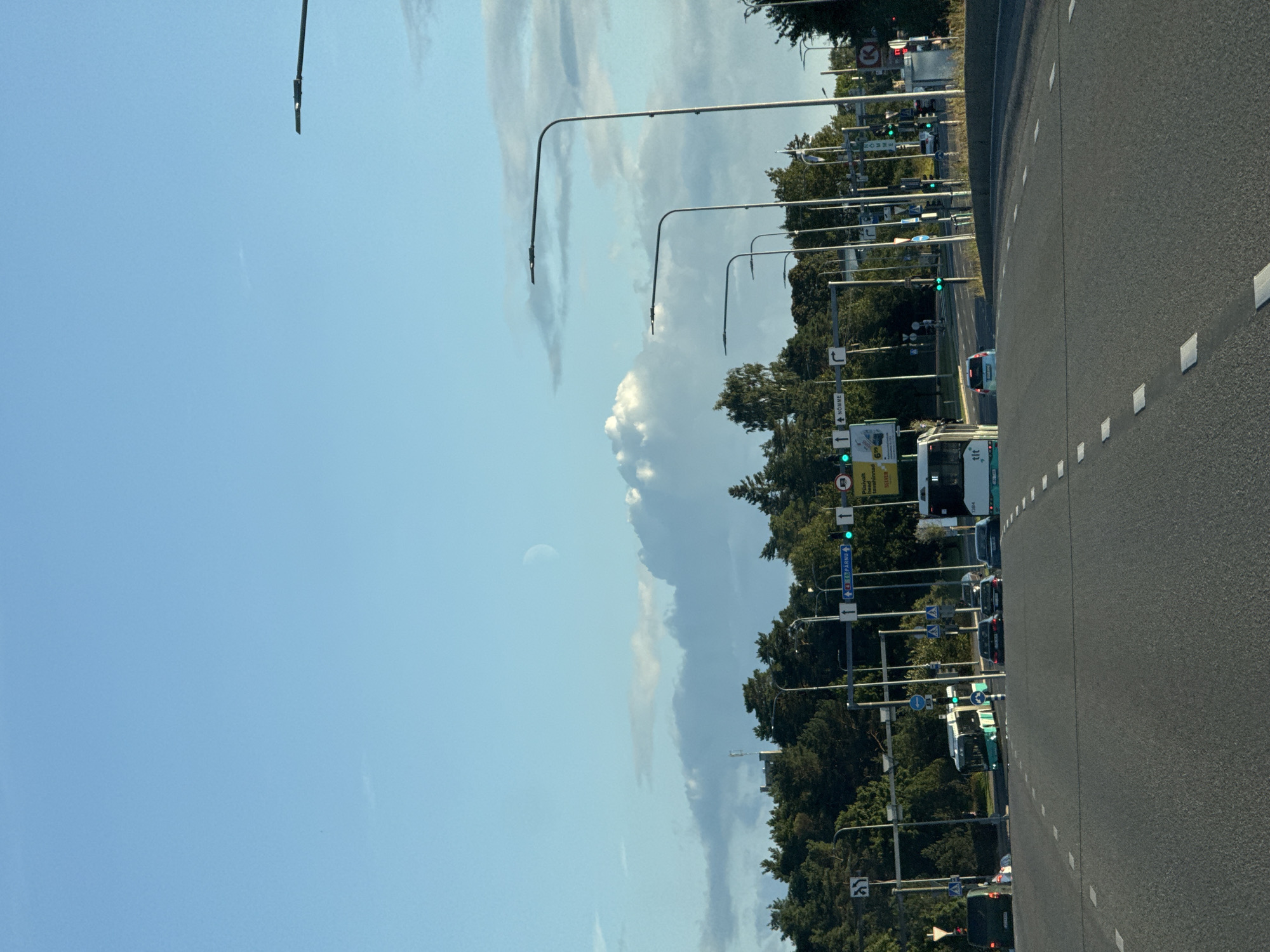 This is a ground-level, eye-level, outdoor, daytime, medium shot of a street scene in Tallinn, Estonia. The image is taken from a moving vehicle on an asphalt road with white lane markings. On the right side of the road, there are multiple streetlights with modern, angled lamp heads and a concrete barrier. Further back, a line of trees forms a dense green border. Traffic lights are visible, some showing green. Several vehicles are on the road, including a bus and cars. There is a billboard on the left with text in Estonian. In the sky, a crescent moon is visible above a large, white cloud formation, with a clear blue sky above. The overall impression is of a typical urban street on a clear day.