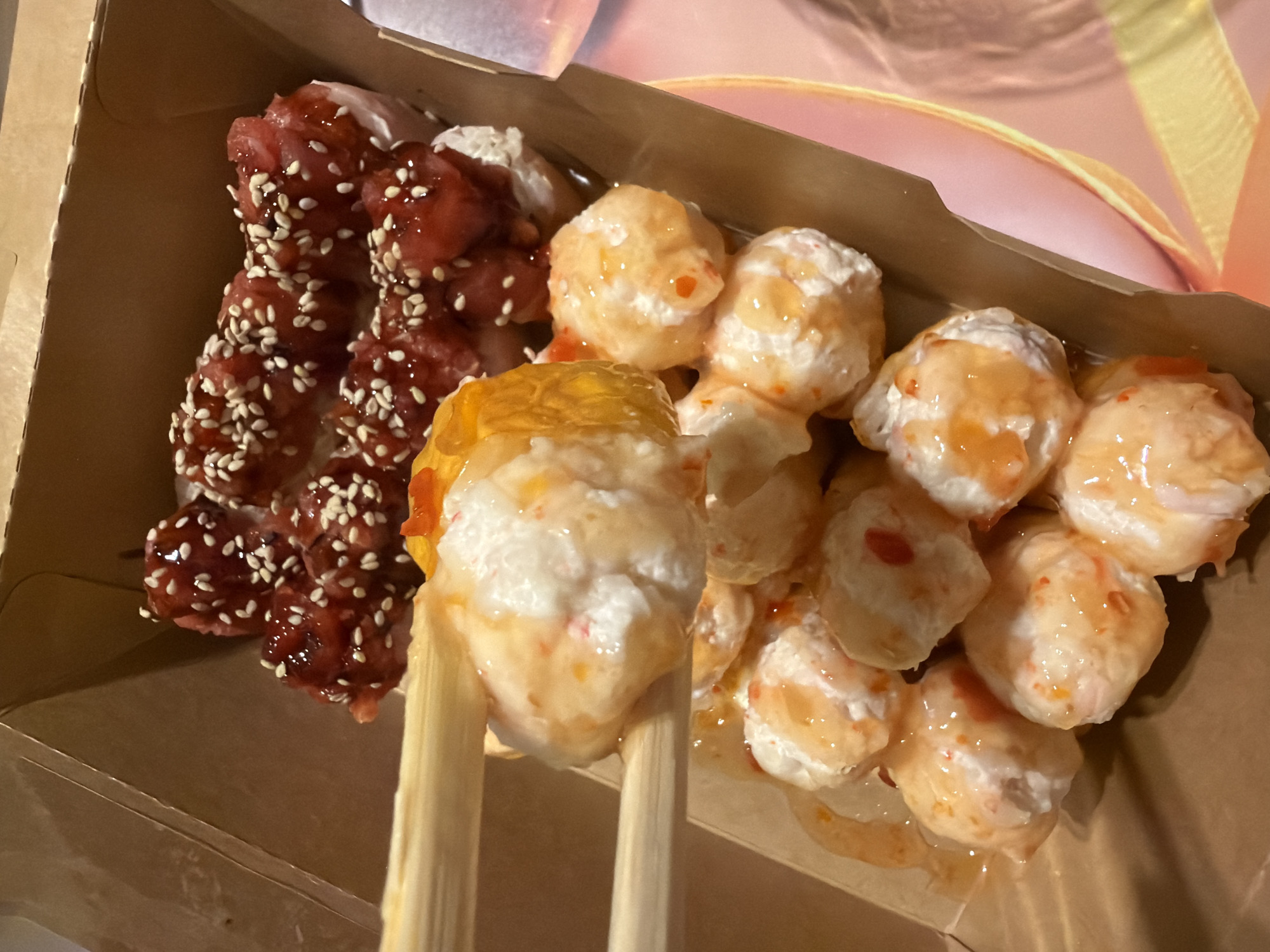 A cardboard container holds two varieties of food items, one of which is being lifted with chopsticks. On the left side of the container is a row of food covered in a red sauce and sesame seeds. To the right is a group of rounded food items coated in a light sauce with small red particles visible within. The chopsticks are positioned in the center of the container, lifting one of the rounded food items. The location in the city of Unknown, Unknown, cannot be confirmed from the image.