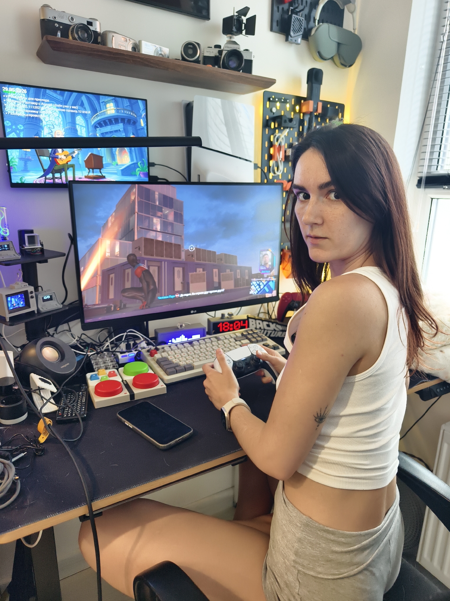 A young woman is sitting at a desk playing a video game. She is wearing a white tank top and gray shorts. She is looking at the viewer with a neutral expression. She is holding a PlayStation controller in her hands.
On the desk in front of her is a keyboard, a phone, two large round buttons (one red and one green), a speaker, and various other electronic devices and cables. A monitor shows the video game "Spider-Man: Miles Morales" being played. Another monitor above it displays what appears to be a cartoon.
Above the monitors is a wooden shelf with several vintage cameras on it. To the right of the monitors, on the wall, are a pair of headphones and some storage panels. A digital clock displays the time as 18:04.
The location is likely indoors, possibly a home office or gaming room. Given the OCR text, there is no indication of this image being in Tyumen, Russia.