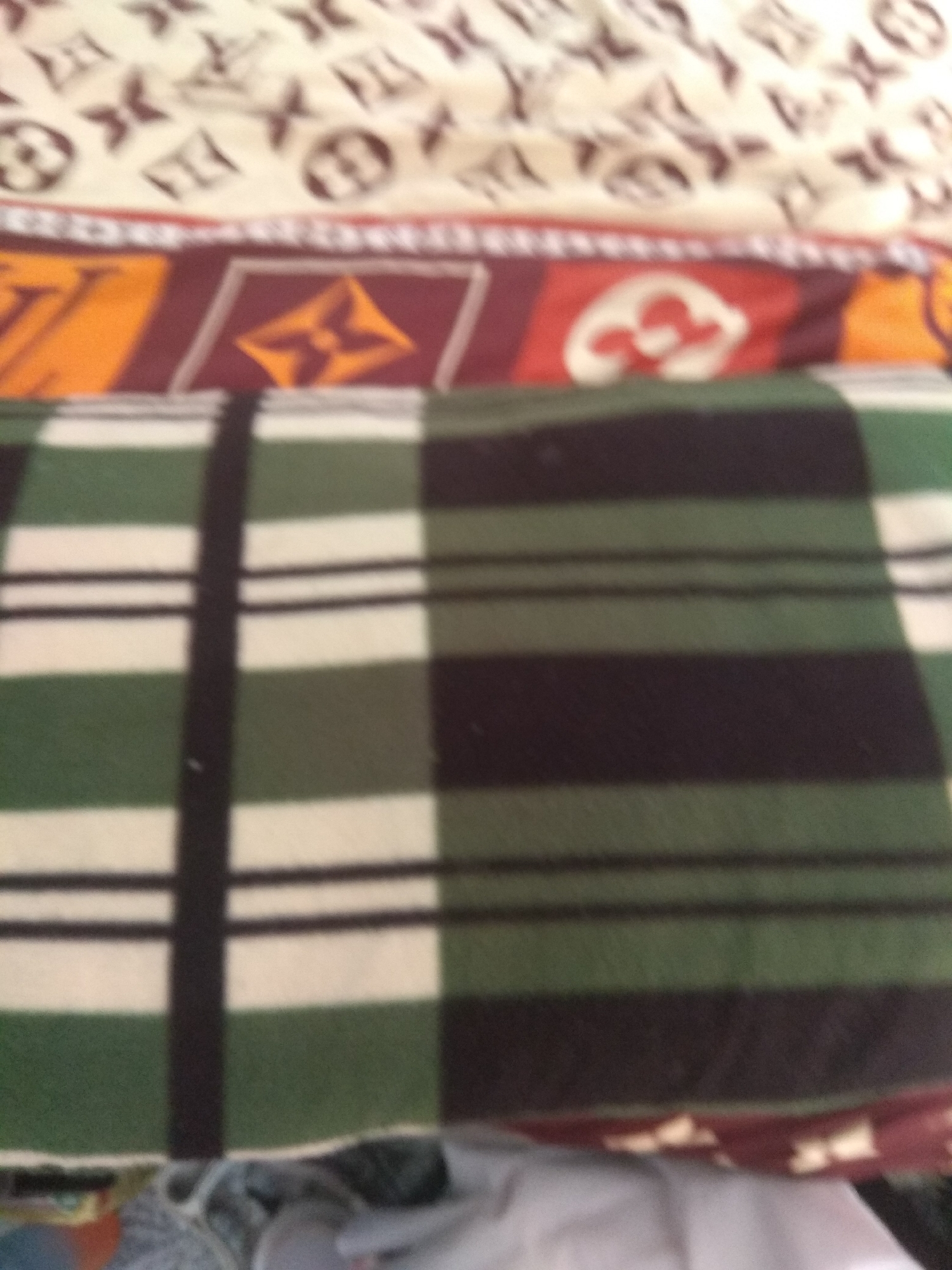 The image shows a close-up of layered textiles. The top layer appears to be a beige fabric with a repeating pattern of brown logos, possibly a luxury brand motif. Below this is a strip of fabric with a maroon border and orange and maroon geometric patterns. The main subject of the image is a folded blanket with a plaid pattern of alternating thick green and black stripes, interspersed with thinner white stripes. The bottom of the image shows a glimpse of other folded fabric items, including a predominantly white material, suggesting a domestic or storage setting. The lighting is dim, and the image is slightly out of focus, obscuring fine details. There are no visible people or any indication of a specific location within Jalingo, Nigeria, or any other city.