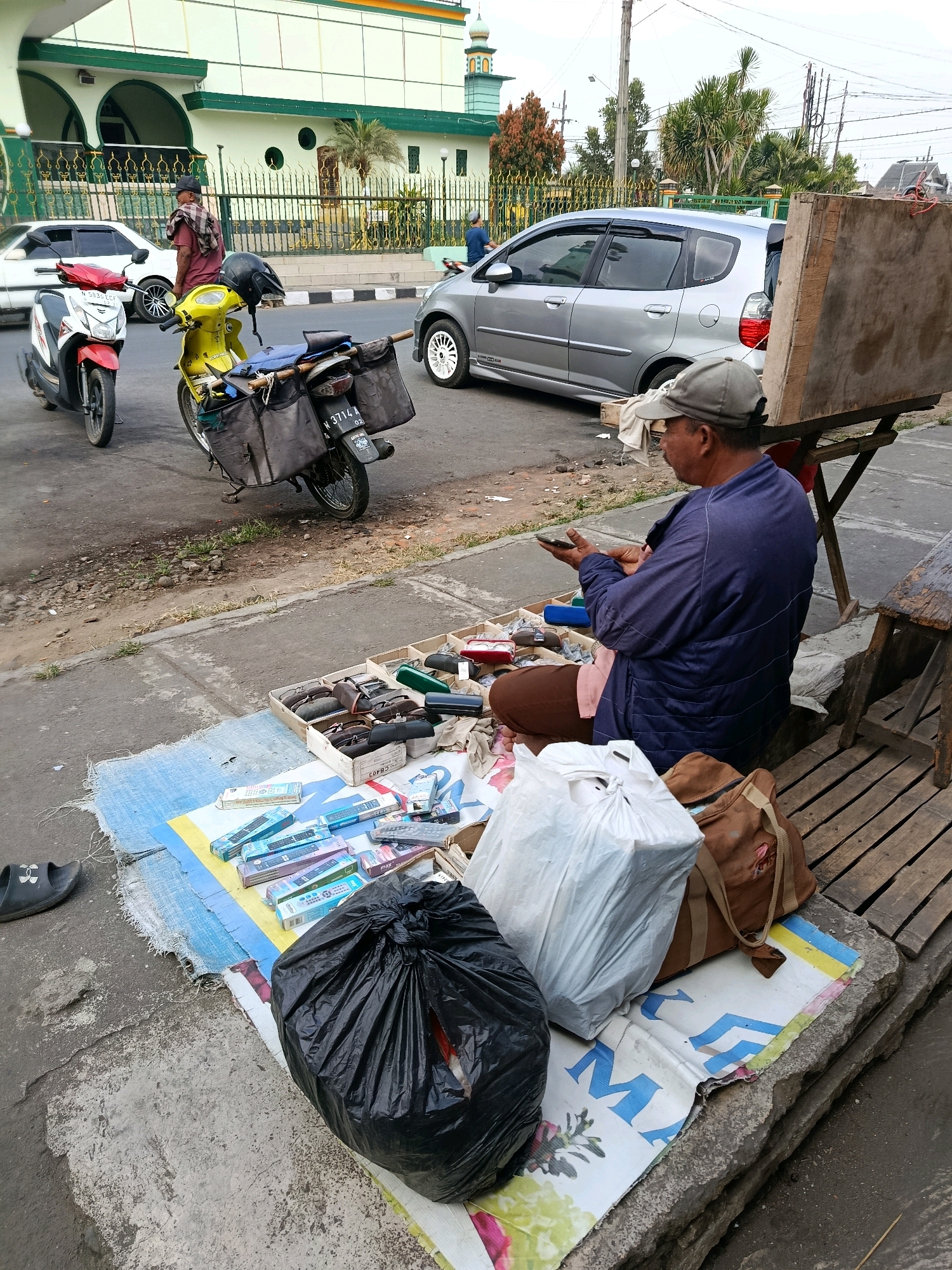 A man sits on the ground next to a pile of goods, including remote controls, and other items displayed on a mat on a sidewalk in front of a mosque in Bululawang, Indonesia. He is wearing a blue jacket, brown pants, and a grey baseball cap. He is holding a mobile phone and appears to be looking at it. Behind him are two motorcycles and a silver car parked on the street. The mosque has a green and white facade with a tall minaret. The environment appears to be a busy street with traffic and pedestrians.