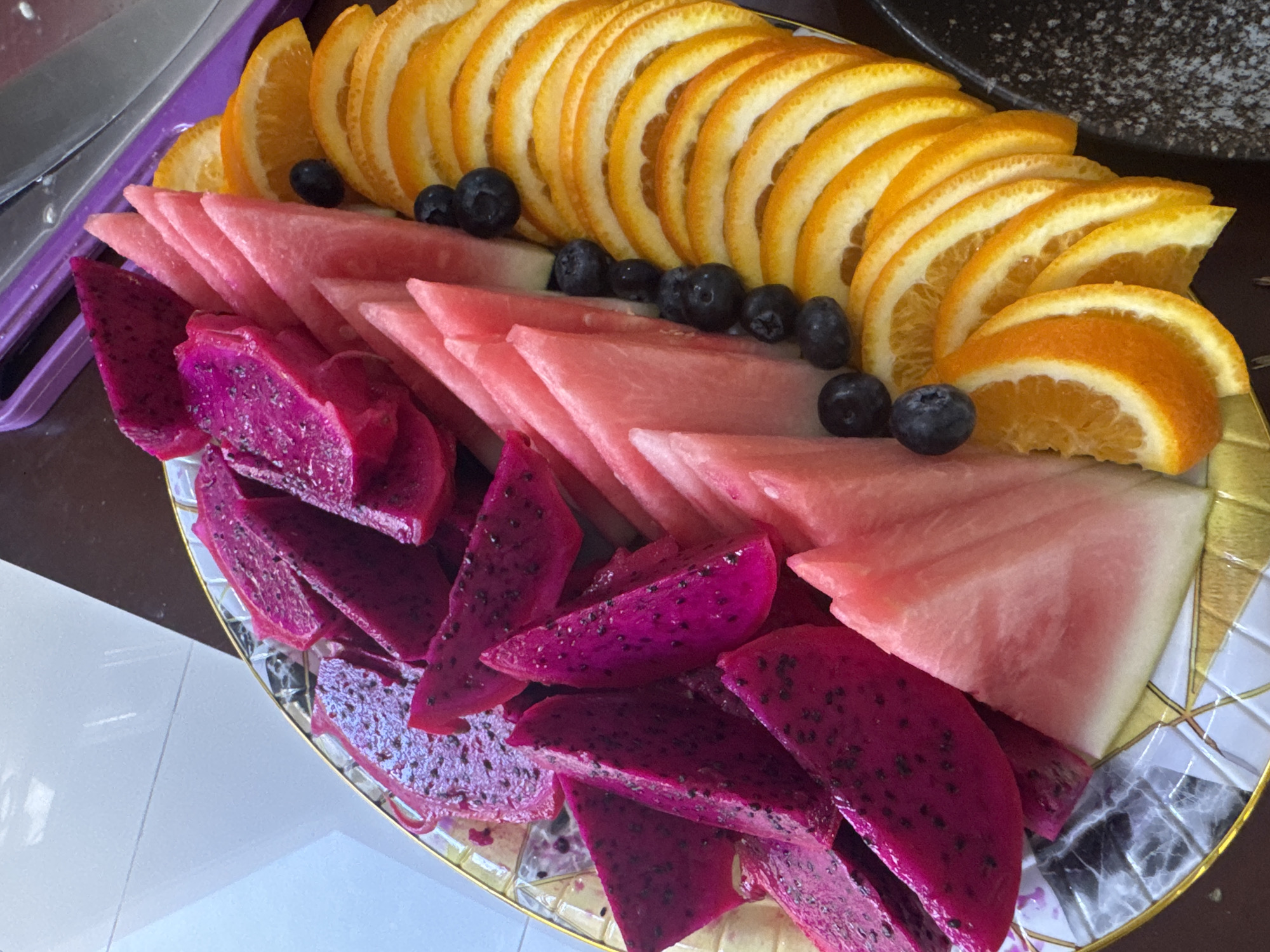 A high-angle, close-up shot depicts a platter of sliced fruits arranged in a decorative pattern. The fruits include orange slices, watermelon wedges, dragon fruit pieces, and blueberries. The orange slices are arranged in a semi-circular row, followed by watermelon and dragon fruit. The blueberries are scattered between the fruit sections. The platter is likely located in Kuta Selatan, Indonesia. No people are visible.