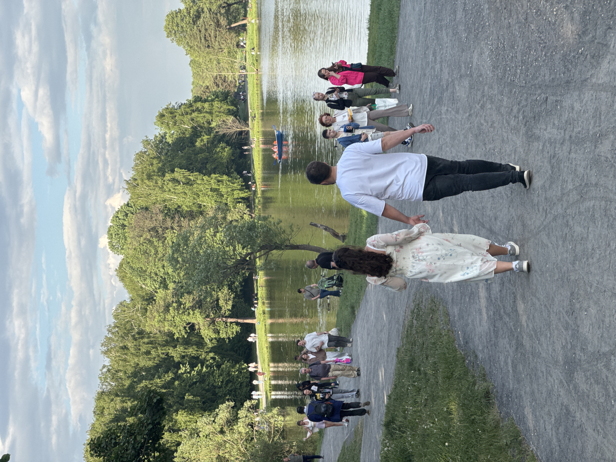 The image depicts a scene in a park, likely in Moscow, Russia. A gravel path runs alongside a body of water, possibly a lake or large pond. On the path, there are several people, including a couple walking hand-in-hand in the foreground. The man is wearing a white shirt and dark pants, while the woman is wearing a light-colored floral dress. Behind them are several other individuals. People are generally casually dressed. The water reflects the surrounding trees and sky, and there are boats on the water in the distance. The sky is overcast with clouds. The environment is lush with trees and greenery.