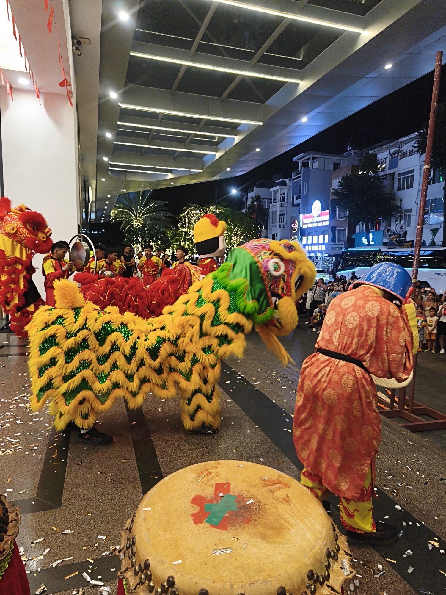 The image depicts a nighttime scene of a street performance, likely a lion dance, in an urban environment. A large, colorful lion costume, predominantly yellow and green with red accents, is the central focus. The lion appears to be operated by two or more individuals beneath it, with their legs visible. In the foreground, a large drum, also yellow with decorative elements, is positioned on the ground. To the right of the drum, a person wearing a red and yellow patterned robe and a blue helmet is visible from the back, appearing to be part of the performance, possibly drumming or directing. Scattered confetti is present on the ground, indicating a celebratory atmosphere.
In the background, several people are gathered, watching the performance. Beyond the crowd, there are buildings with illuminated windows, suggesting commercial establishments or residences. Palm trees and string lights are also visible, adding to the festive ambiance. The overall lighting suggests artificial illumination from streetlights and building lights. The location is identified as Unknown, Unknown.