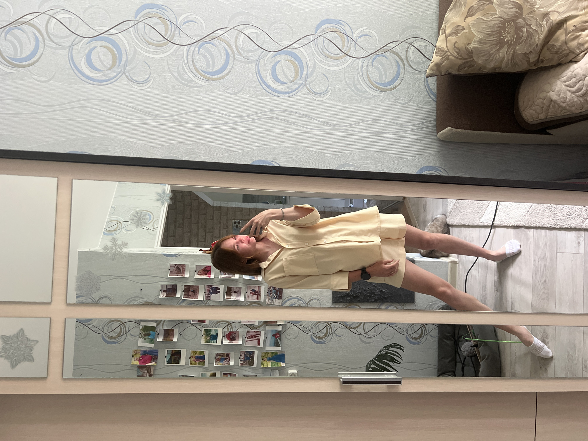 The image shows a woman taking a photo of herself in a mirror. The woman is wearing a light yellow pajama set. Her hair is brown, and she is wearing a red hair accessory. She is holding a smartphone to take the photo. The mirror reflects a room with wallpaper, photos attached to the mirror, and a sofa. A cat is visible near the woman's feet. The photo was taken in Chaikovsky, Russia.