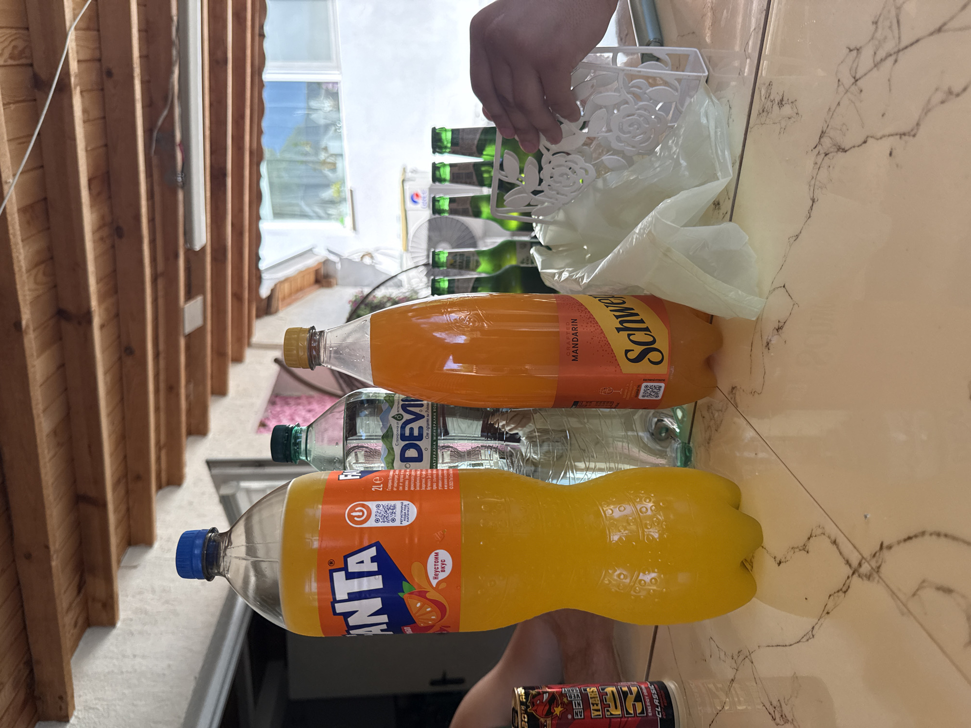 This indoor shot depicts a collection of beverages on a tiled surface. From top to bottom, there's a hand holding a white, floral-patterned container, followed by multiple green-bottled drinks, a bottle of Schweppes Mandarin, a bottle of "Devin" mineral water, and a bottle of Fanta. A can of energy drink is also present. The setting seems to be in Elhovo, Bulgaria based on the Bulgarian text on the Fanta bottle. A partially visible wooden structure and exterior landscape are in the background, providing depth to the scene.