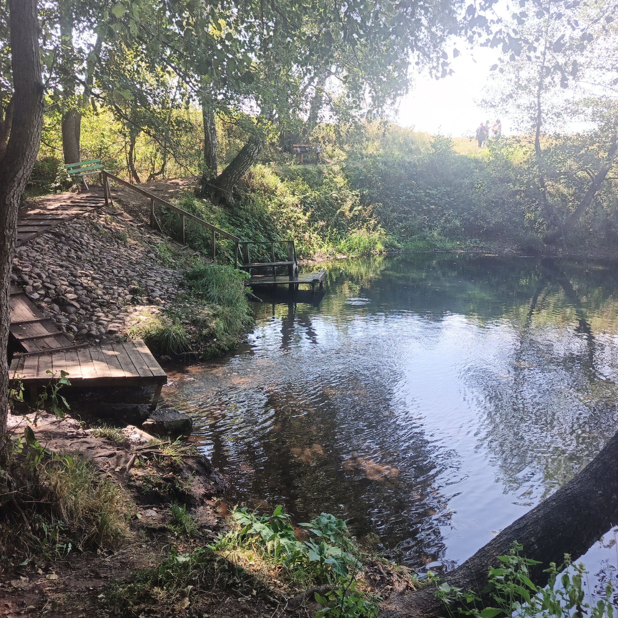 The image depicts a natural outdoor scene near a body of water, possibly a spring or a pond, located in Tugustemir, Russia. The foreground features a wooden platform and steps leading down to the water's edge, with a pathway composed of cobblestones and wooden planks ascending a slope on the left. Dense green foliage and trees surround the area, casting dappled shadows. The water itself is calm with reflections of the trees and sky visible on its surface. On the opposite bank, more vegetation and trees line the water. In the distance, on a sunlit grassy hill, two figures are visible, appearing as small silhouettes against the bright sky. A wooden bench is situated on the pathway near the top of the slope. The lighting suggests it is daytime, with sunlight filtering through the tree canopy.