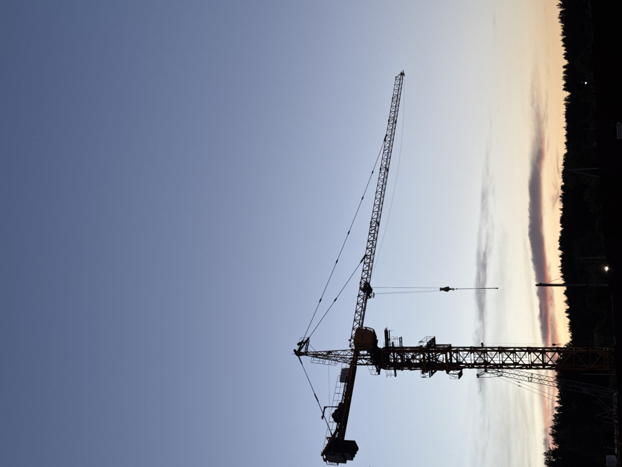 The image shows a construction crane against a sky that transitions from blue to sunset colors. The crane is silhouetted, obscuring details, but its boom and support structure are visible. To the right, a dark line of trees suggests a treeline or forest edge. The image is rotated counterclockwise. There is no evidence of the location being Izhevsk, Russia, nor any people, subjects, objects, actions, interactions, or environment elements to support this claim.