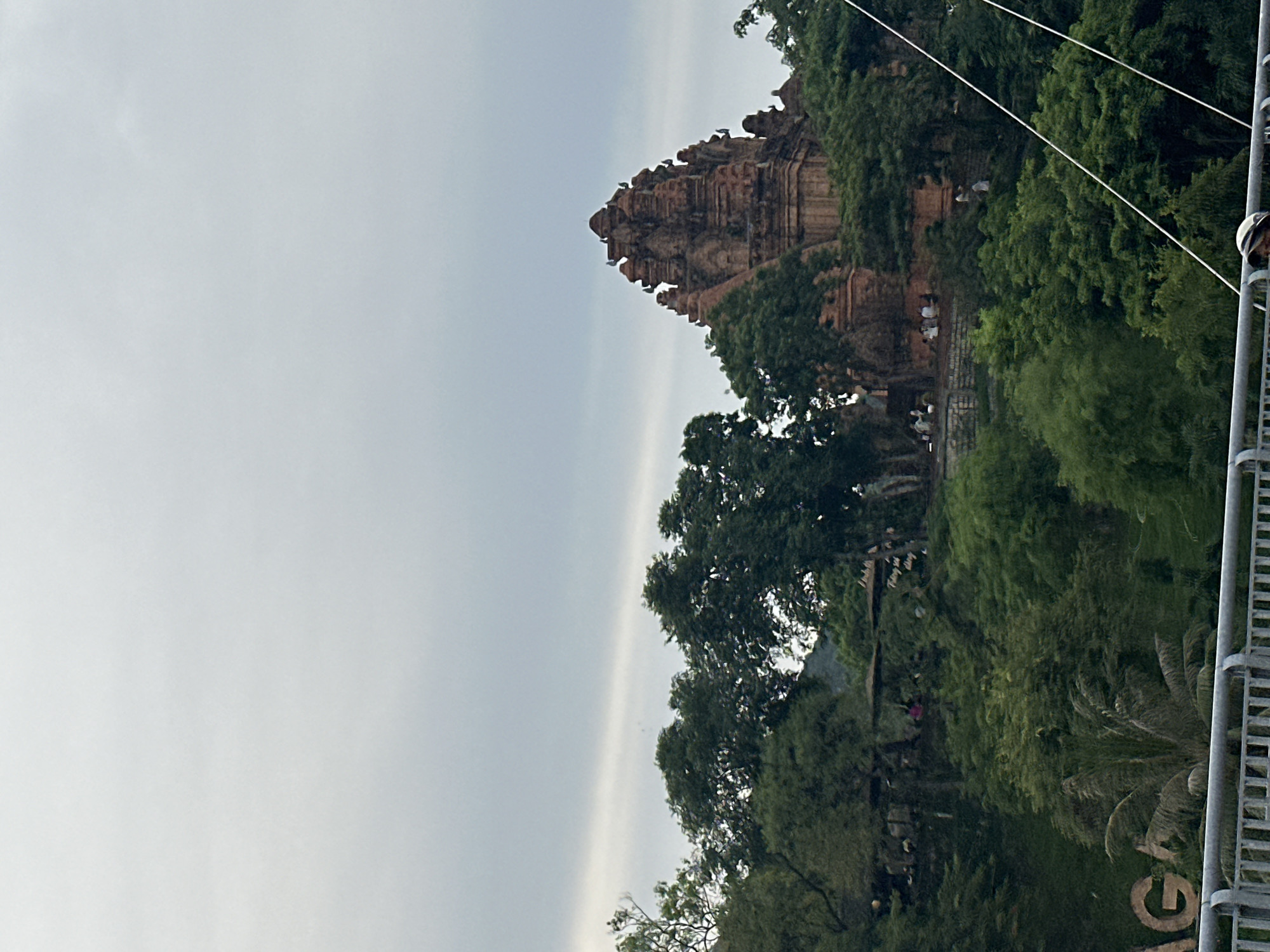 This is a medium shot, eye-level, exterior, daytime, vertical, full shot of the Po Nagar Cham Towers in Nha Trang, Vietnam. The towers are made of red brick and are nestled within a lush green landscape. The sky is overcast and grey. In the foreground, a metal railing is visible, and a person wearing a white helmet is partially visible in the bottom right corner. The towers themselves are multi-tiered and appear to be ancient structures, with intricate carvings and architectural details. The surrounding trees are dense and varied in shape and size, creating a natural canopy around the historical site. There are small groups of people visible on the terraces leading up to the towers, some appearing to be seated. The overall impression is one of a serene and historical place of worship.