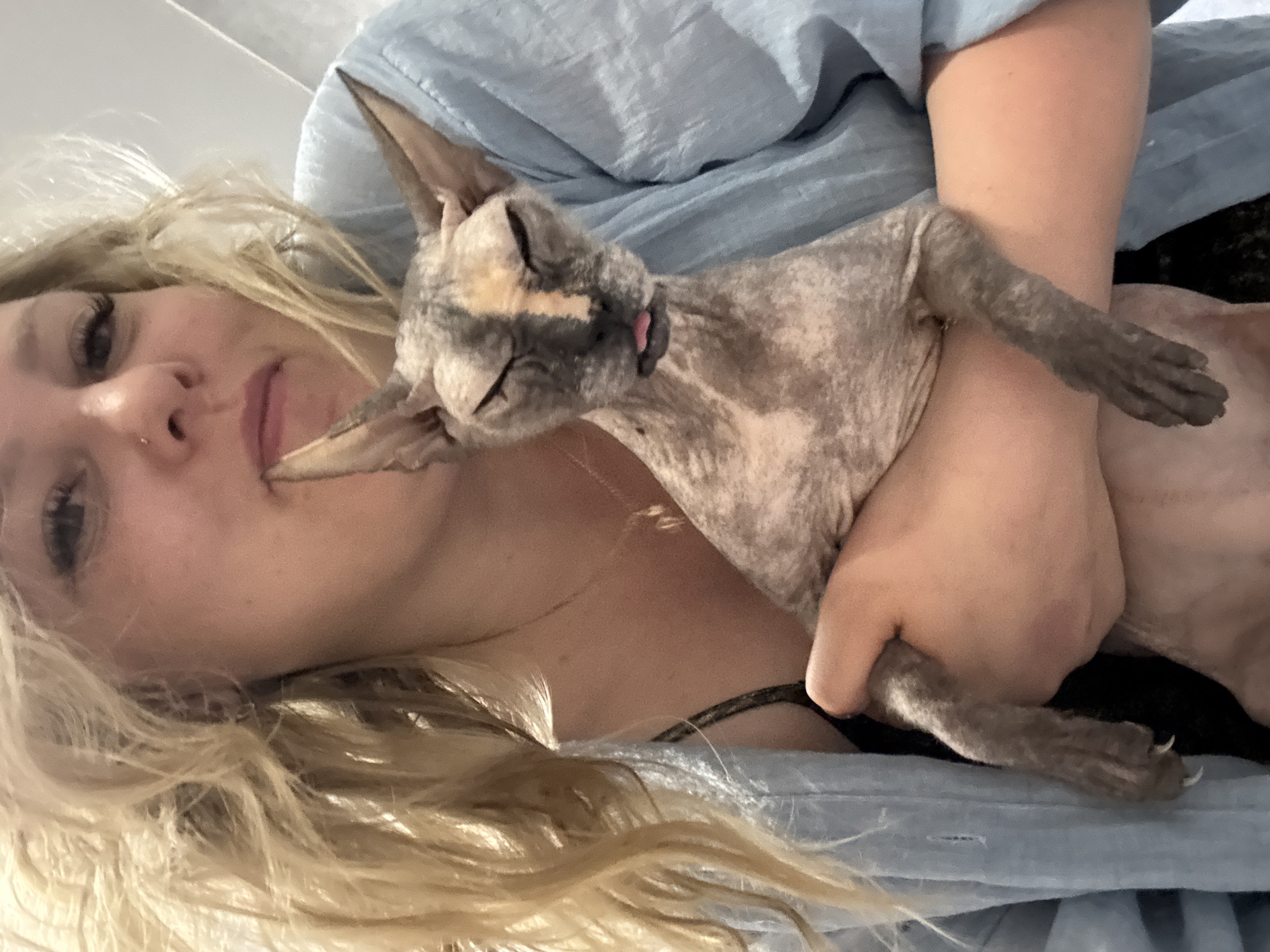 A woman with long blonde hair is lying on her side, holding a grey and beige Sphynx cat. The cat is lying on its back in her arms, with its eyes closed and tongue slightly sticking out. The woman is wearing a light blue shirt. The location cannot be determined from the image.