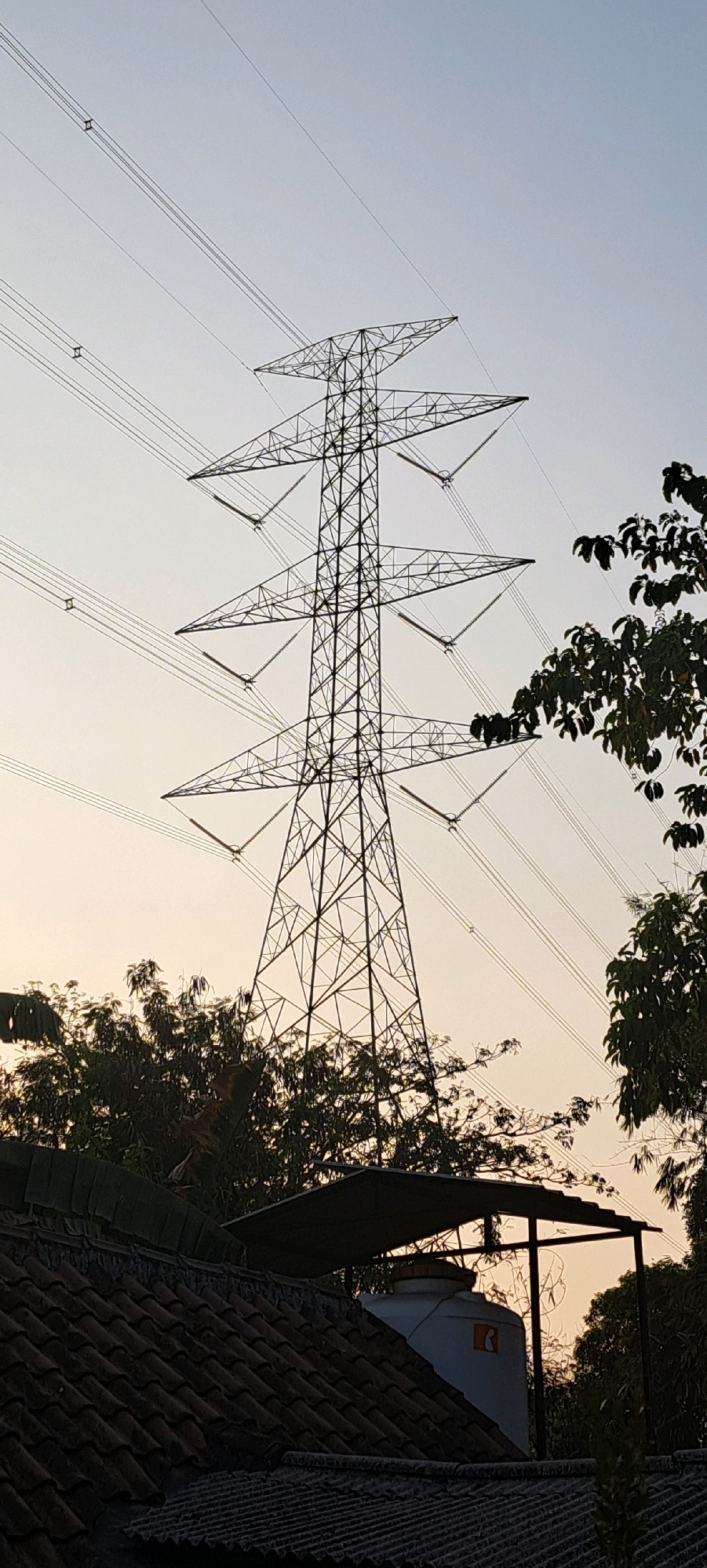 The image shows a towering electricity pylon in Kemiri Sewu, Indonesia, silhouetted against a pale sky, likely at dawn or dusk. The pylon is made of a lattice of metal beams, with multiple sets of high-voltage power lines extending from its arms. In the foreground, the roof of a building with traditional red tiles is visible, along with a water tank partially covered by a metal canopy. Lush green trees and foliage surround the structure, partially obscuring the lower parts of the pylon. The scene depicts a blend of industrial infrastructure and natural elements within a residential or semi-rural setting.