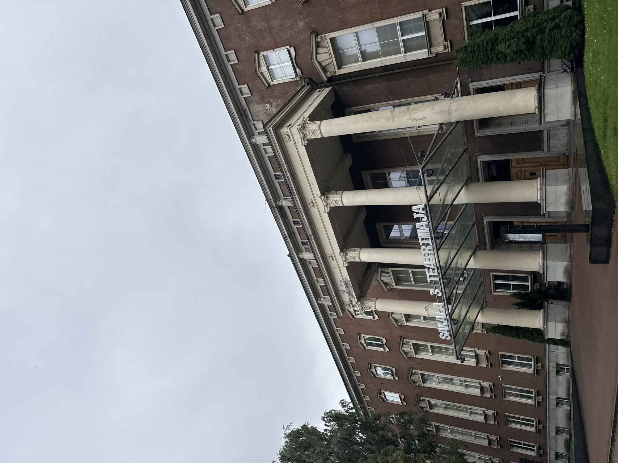 The image shows the exterior of a building with a maroon-colored facade, featuring a classical architectural style with large columns supporting an overhang. The building has numerous tall, rectangular windows with white frames. A glass canopy extends over the entrance, and white text is visible on it, reading "SAKALA 3 TEATRI MAJA". The sky is overcast and grey. To the left of the building, a tall, leafy tree is partially visible, and to the right, a few evergreen trees are planted. The foreground consists of a red brick walkway leading to the building, with a patch of green grass to the right. The building appears to be a public institution, possibly a theater or cultural center, located at Sakala 3 in Tallinn, Estonia. No people are visible in the image.