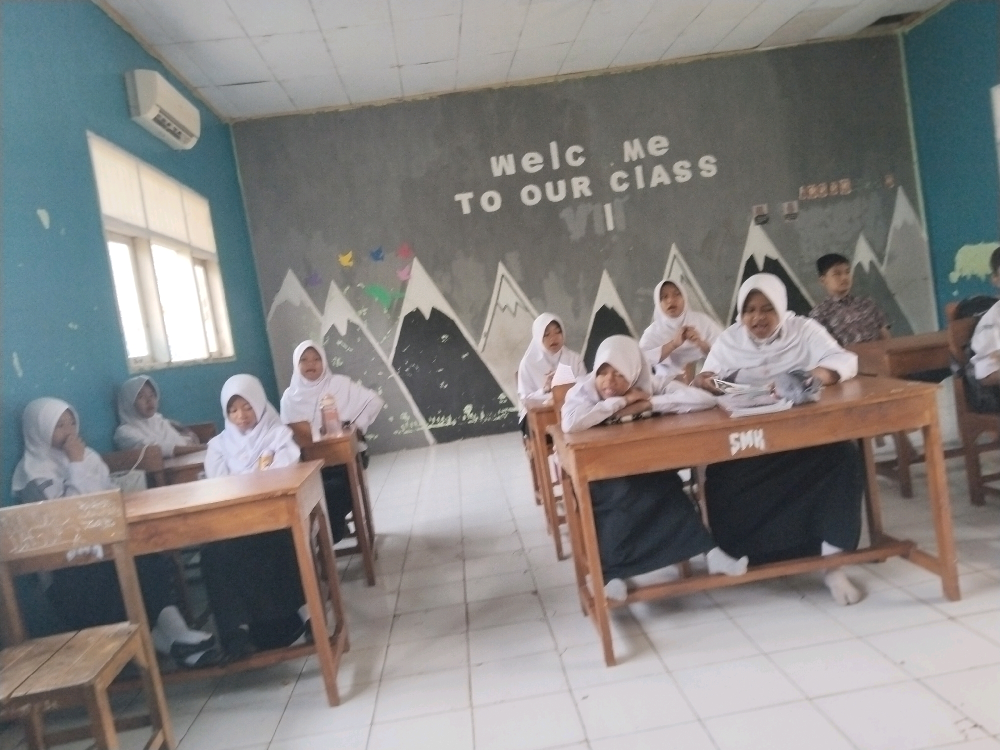 The image depicts an indoor classroom scene, likely in Serang, Indonesia, featuring young students, predominantly girls, wearing white hijabs and uniforms. They are seated at wooden desks in what appears to be a school setting. The classroom walls are painted in blue and gray, with the gray section featuring a mural of mountains and the text "WELCOME TO OUR CLASS VII" painted in white. The floor is tiled in a white checkered pattern. Several students are visible in the foreground and mid-ground, some appearing engaged in activities like reading or writing, while others look towards the camera or rest their heads on their desks. The presence of a whiteboard with some faint writing, an air conditioning unit mounted on the wall, and school desks suggests a functional educational environment. The overall atmosphere appears to be that of a typical school day, with students participating in classroom activities.
