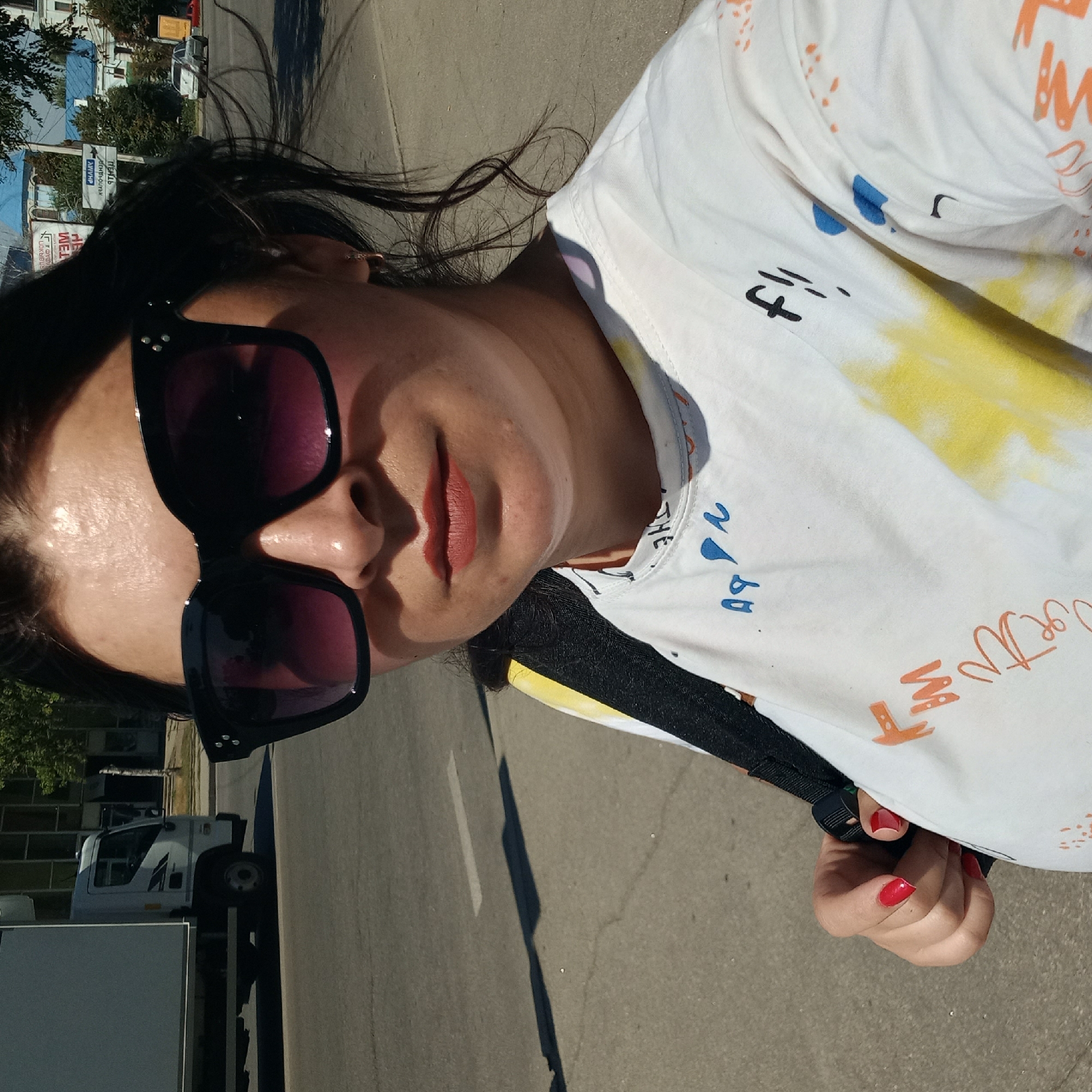 A woman with dark hair, wearing large black sunglasses, a white t-shirt with colorful abstract designs, and red nail polish, stands on a paved street in Kropyvnytskyi, Ukraine. She is holding a black strap with her right hand. In the background, a white truck is parked on the street, and buildings and trees are visible. The sky is bright and sunny.