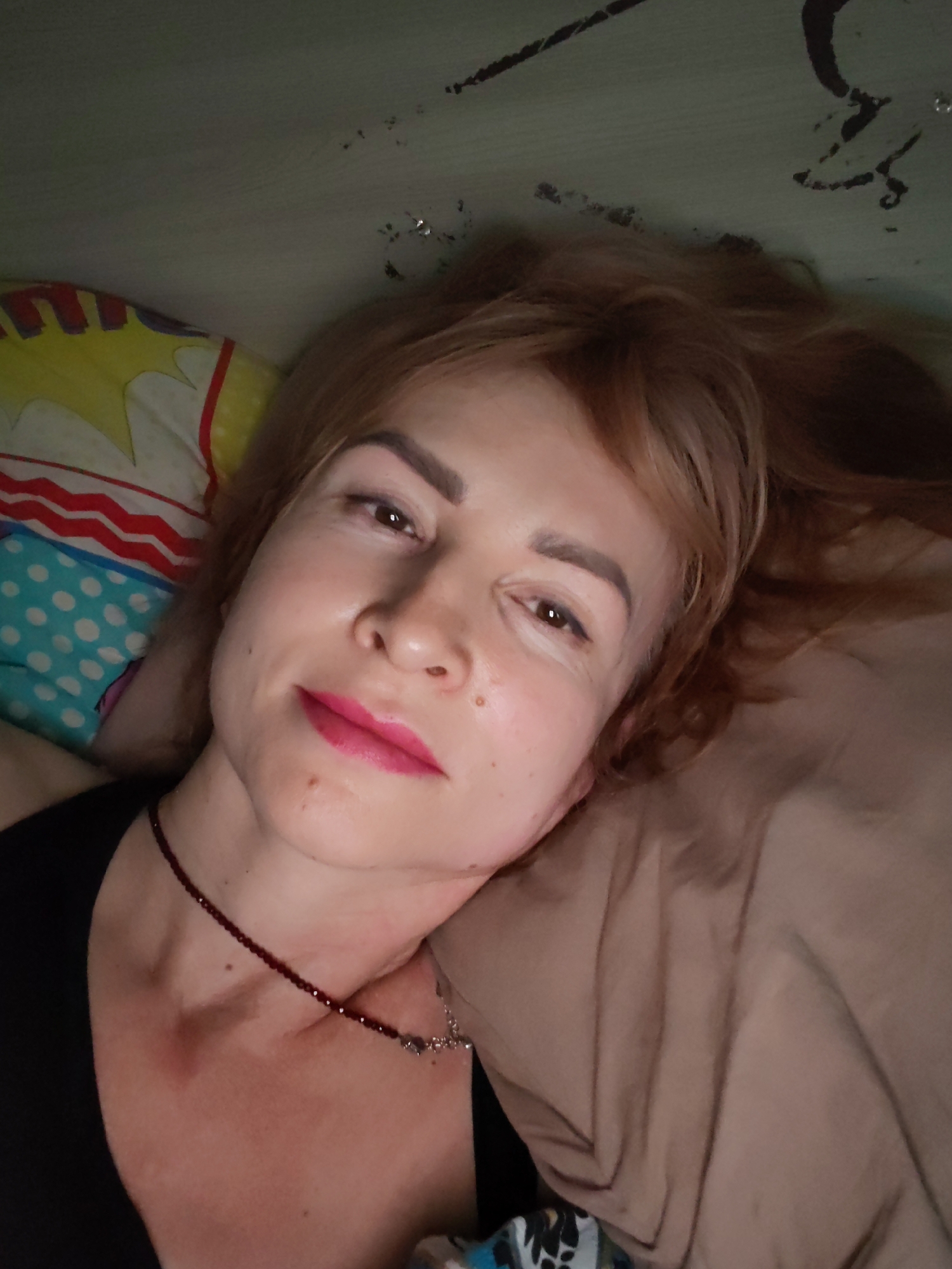 A woman with reddish-blonde hair is lying on a bed. She is facing the camera. She is wearing a black top and a maroon beaded necklace. Her lips are colored with pink lipstick. She is resting her head on a tan pillow. A colorful pillow with comic book style designs and polka dots is visible next to her. The background appears to be a wall with some dark markings. The image was taken indoors. There is no mention of the city of Chaikovsky, Russia.